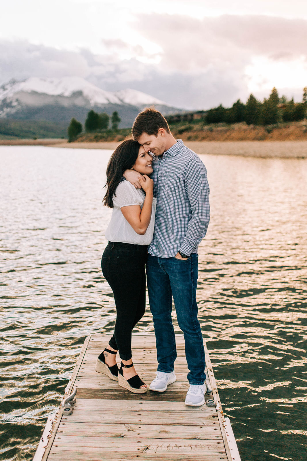 Couple snuggling during engagement photos while standing on lake dock with mountain backdrop in Breckenridge Colorado