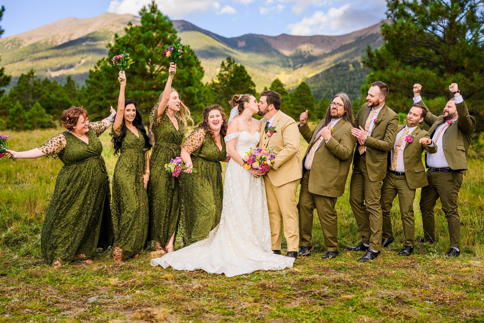 bride and groom kissing while bridal party cheers and smiles in Flagstaff with mountain background and pine trees