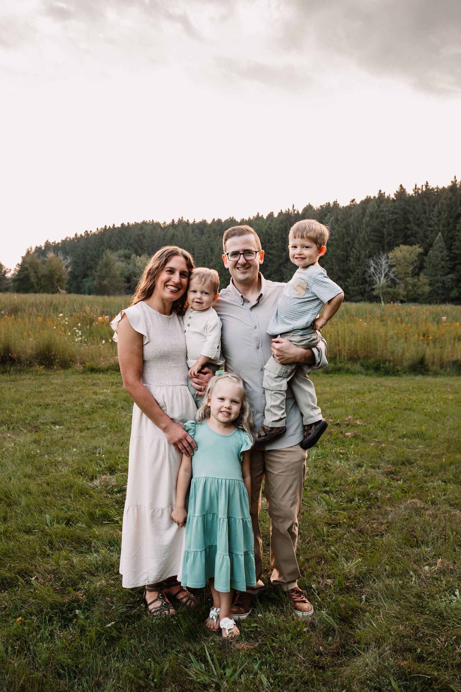 A mom and dad hold 2 toddlers with their oldest daughter standing with them in a field at sunset