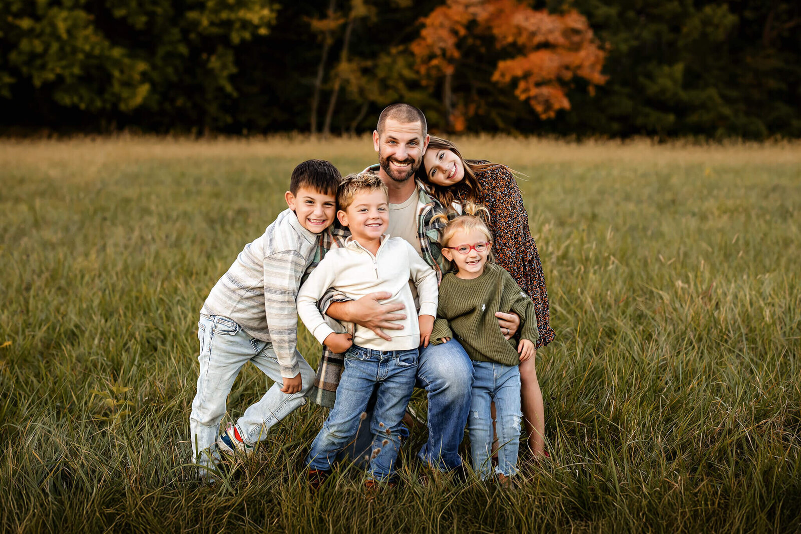 A man and four children stand closely together, smiling in a grassy field. The background features trees with green and orange leaves, suggesting an autumn setting. Captured by a Bloomington family photographer, the group is casually dressed and appears joyful and relaxed in this outdoor portrait.