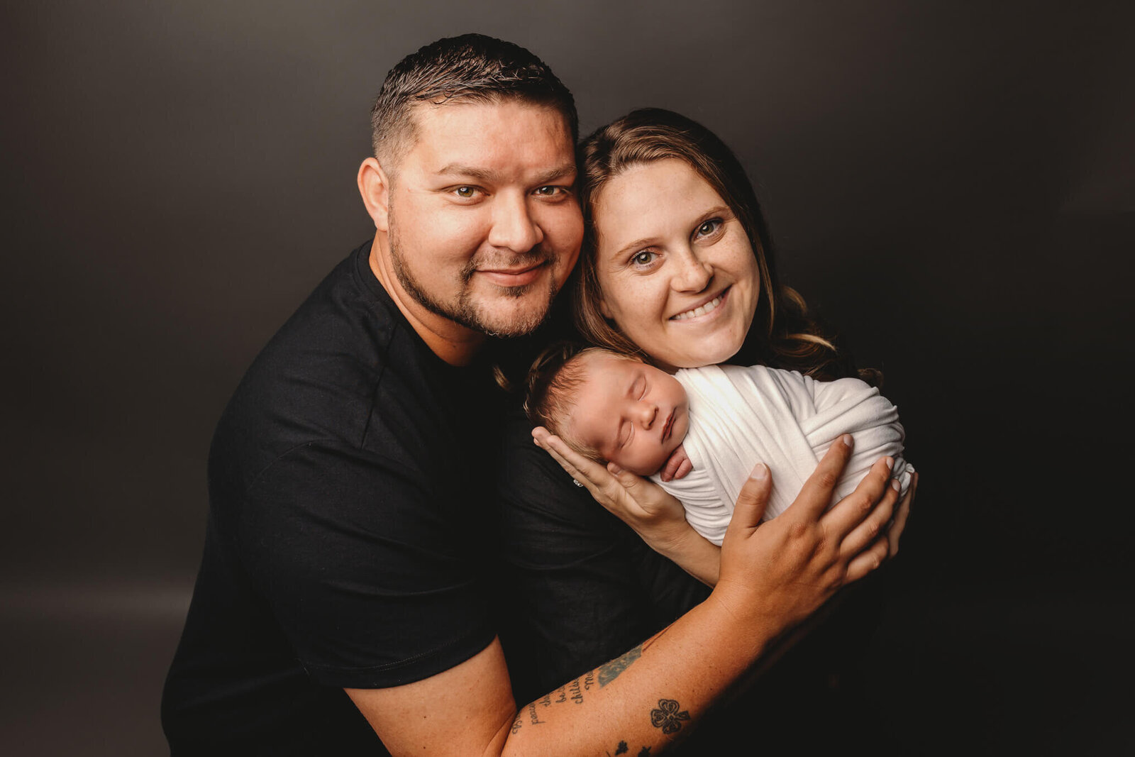 Mom and dad smiling and holding swaddled newborn baby in studio newborn session