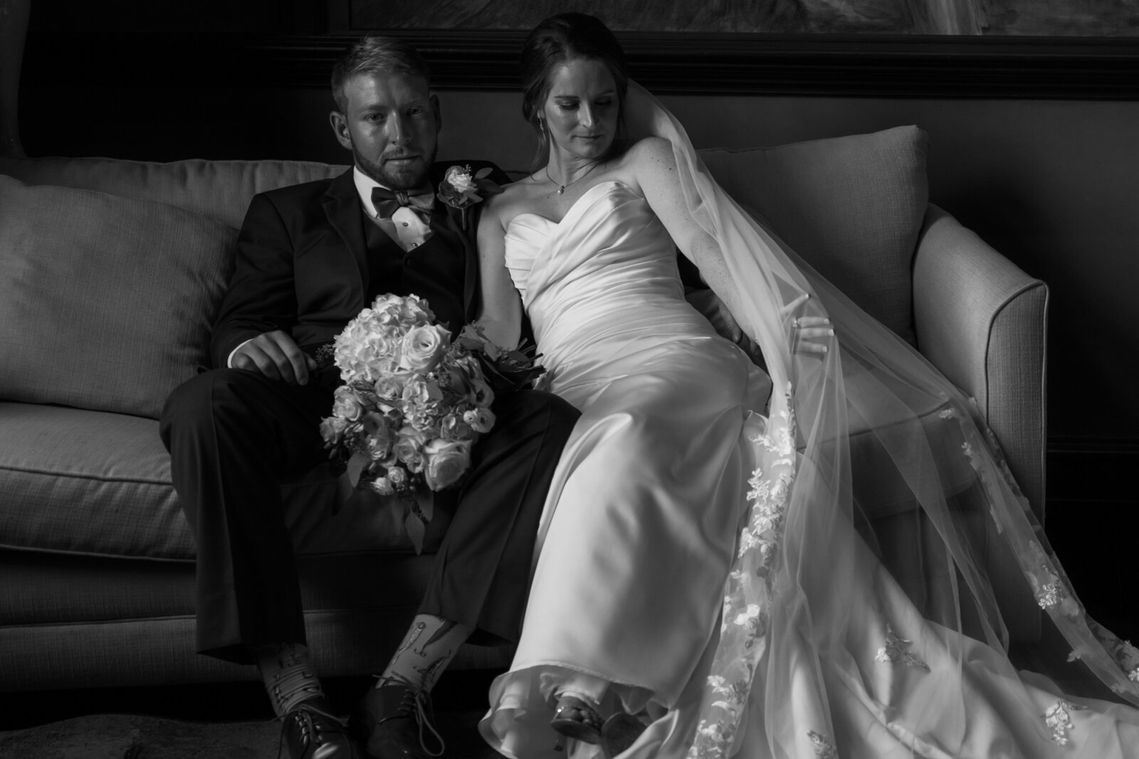 A bride with a long veil sits with a groom on a couch