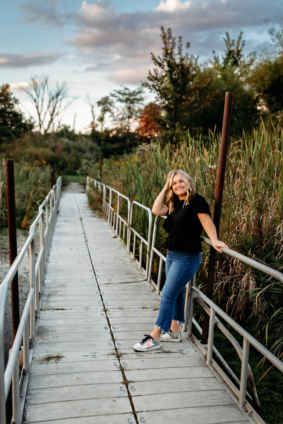 A person with long blonde hair stands smiling on a wooden walkway amidst lush greenery. They rest one hand on the railing and wear a black shirt, blue jeans, and white sneakers. The sky is partly cloudy, hinting at sunset.