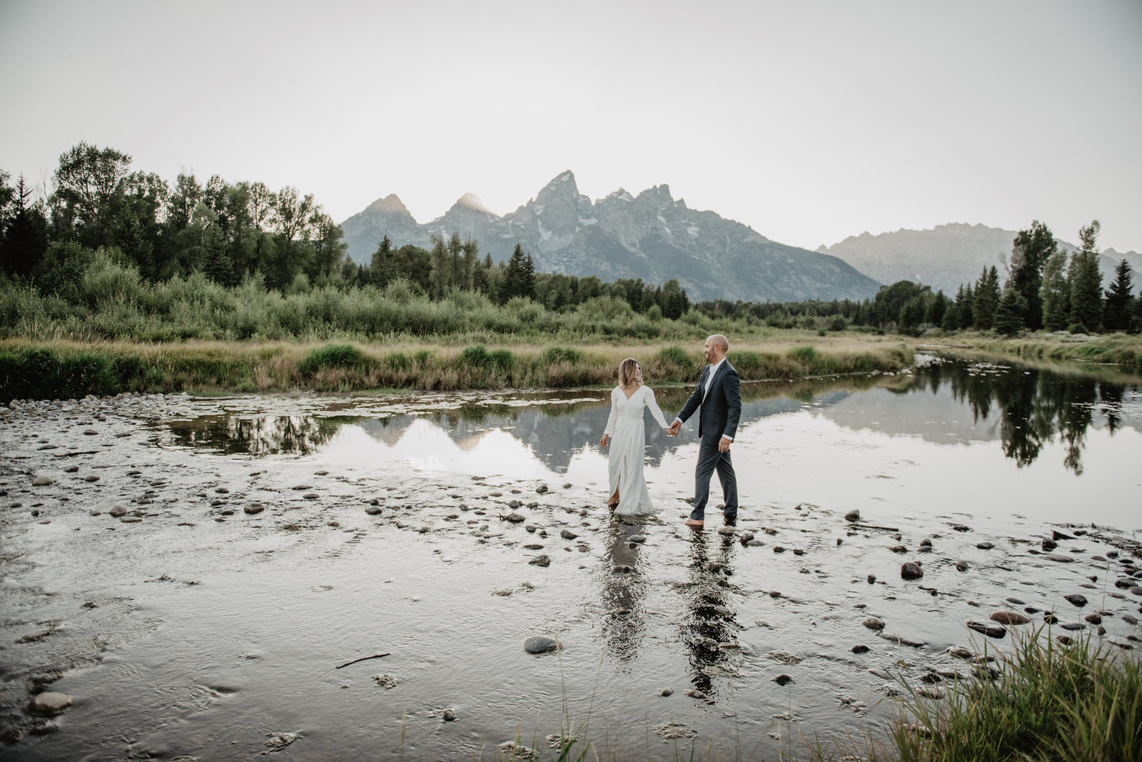Grand Teton wedding with bride and groom holding hands as they cross the snake river together with the Grand Tetons in the background photographed by Jackson hole wedding photographer