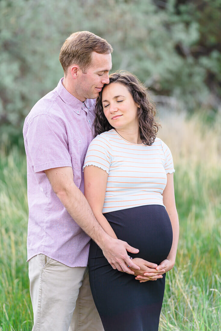 A husband wearing a pink button down shirt and khaki pants holds his pregnant wife wearing a white striped shirt and black skirt as they stand in the tall grass at North Lake Park in Lehi. Captured by Utah maternity photographer Melissa woodruff photography