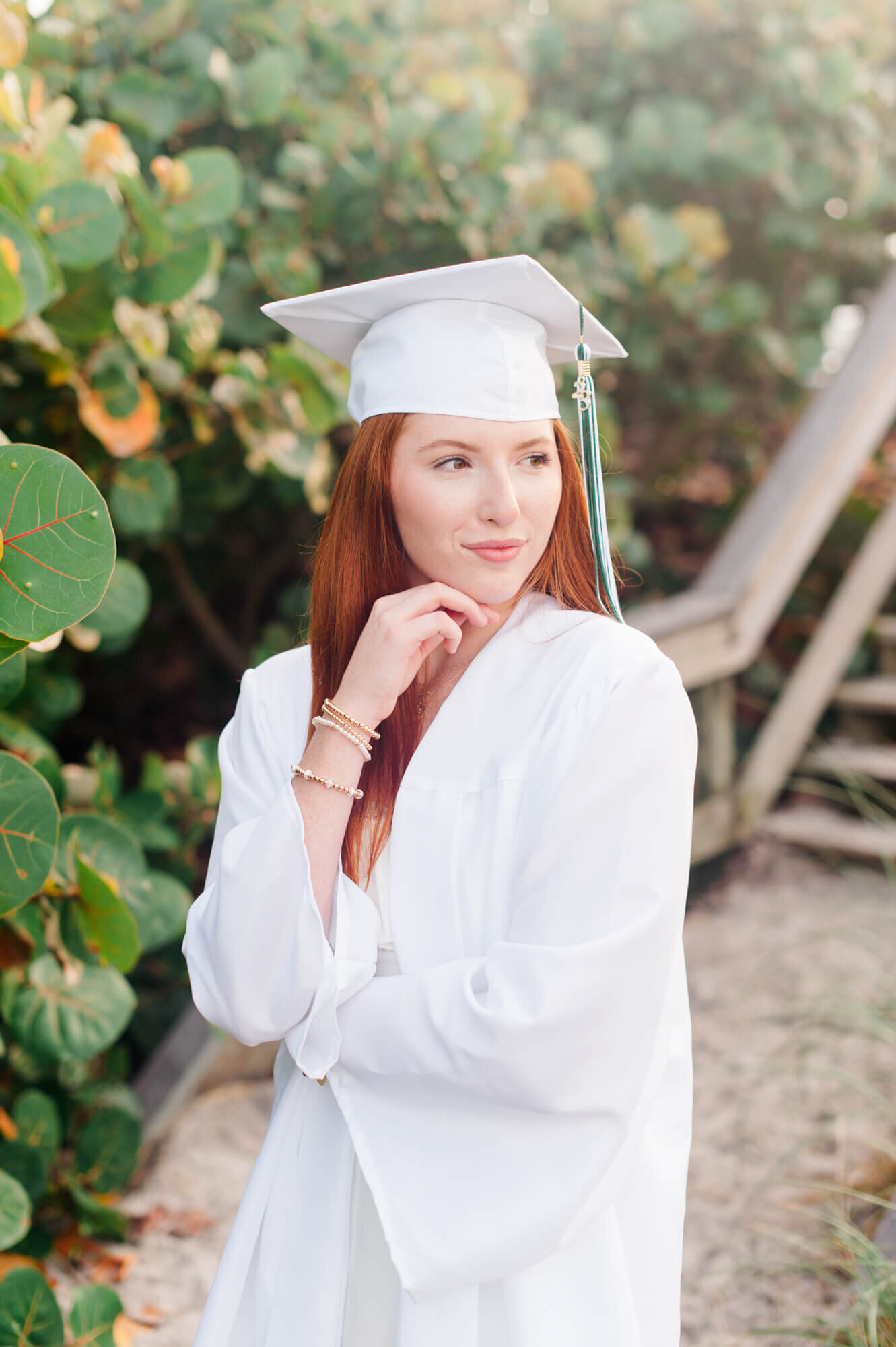 Senior stands near boardwalk during her senior session wearing aa white cap and gown