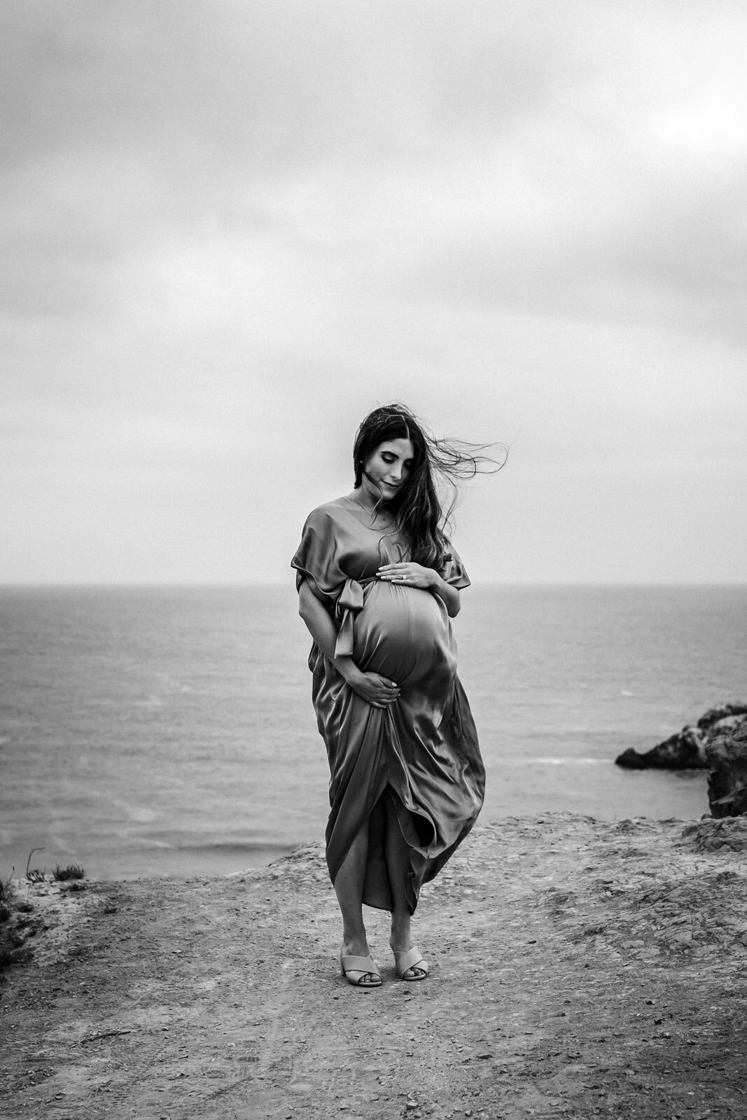 pregnant woman standing above pacific ocean in san franisco holding baby bump with wind blowing her hair and dress