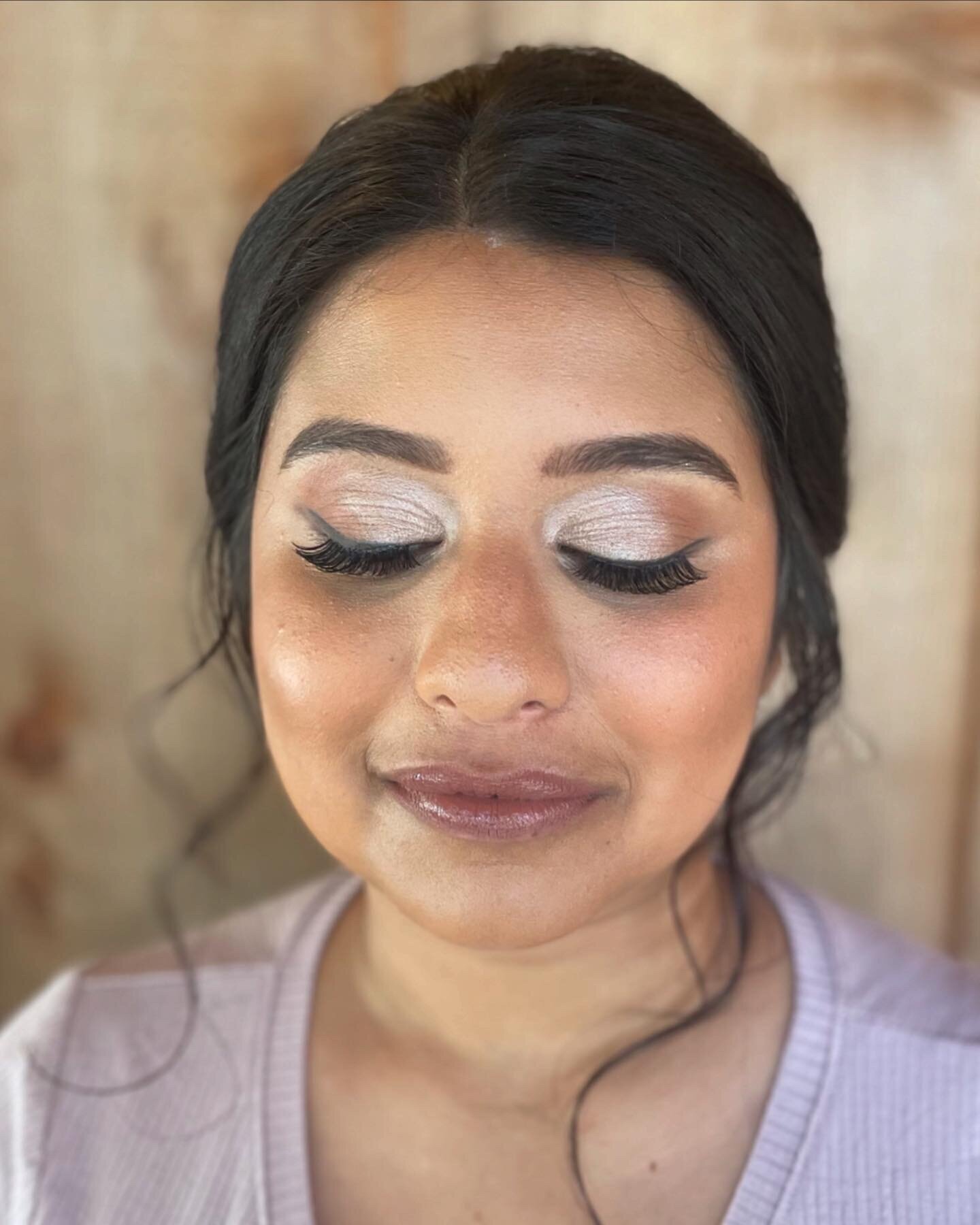 Woman with closed eyes and long lashes, showcasing natural glam wedding makeup while wearing a light purple top against a wooden background.
