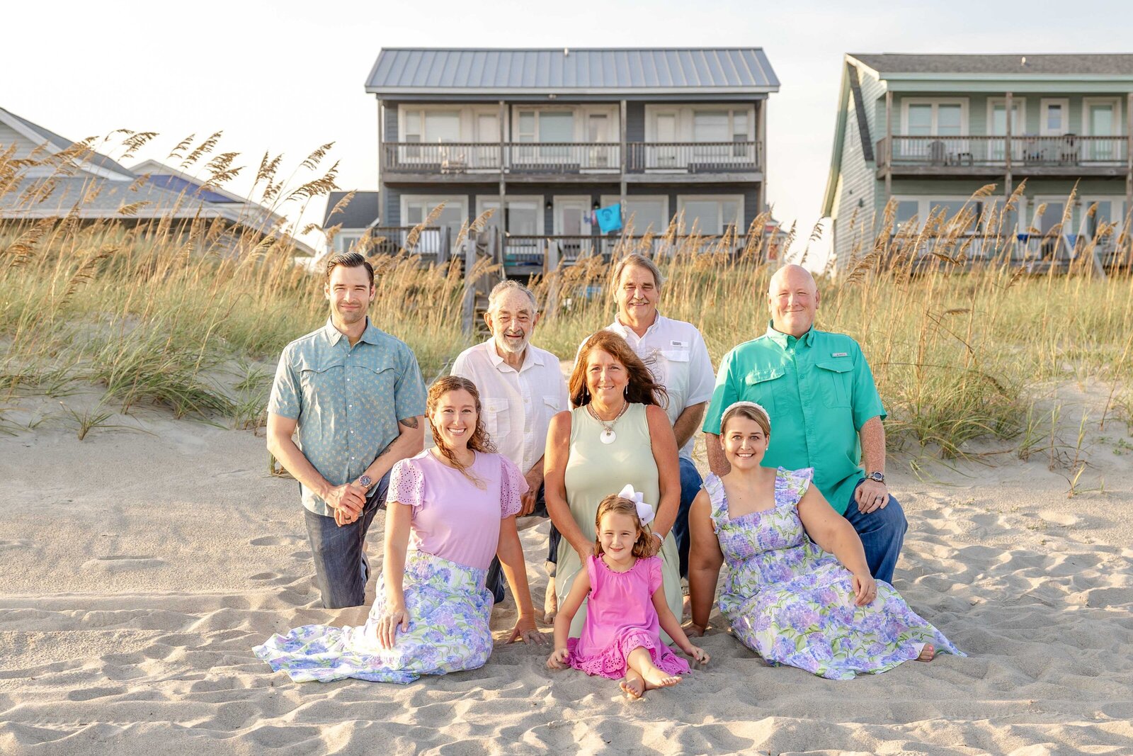 A family poses on the sandy beach with coastal homes and sea oats in the background. Dressed in pastel and floral outfits, they enjoy a sunny day, ideal for destination family photographer NC, highlighting relaxed and joyful beachside moments.
