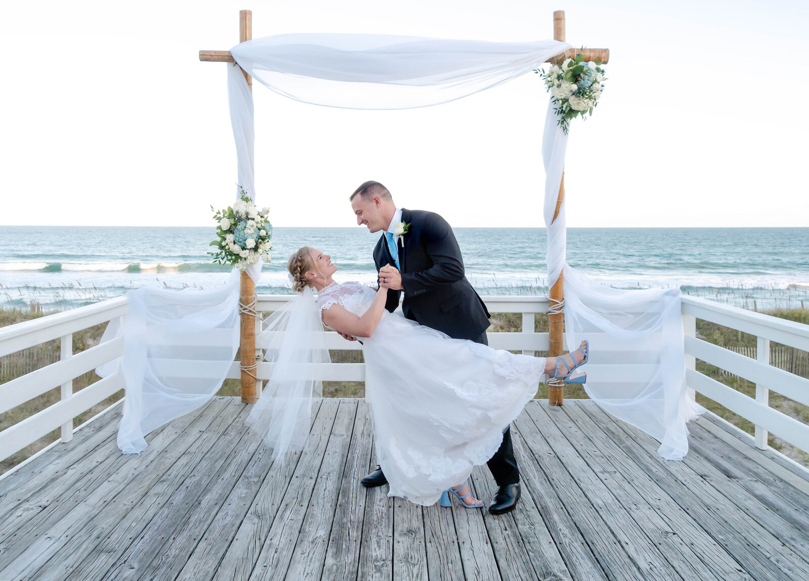 A groom dips his bride on an oceanfront deck under a rustic ceremony arch adorned with flowing white fabric and floral arrangements, captured during a Topsail Beach, NC wedding, blending coastal charm and elegance.