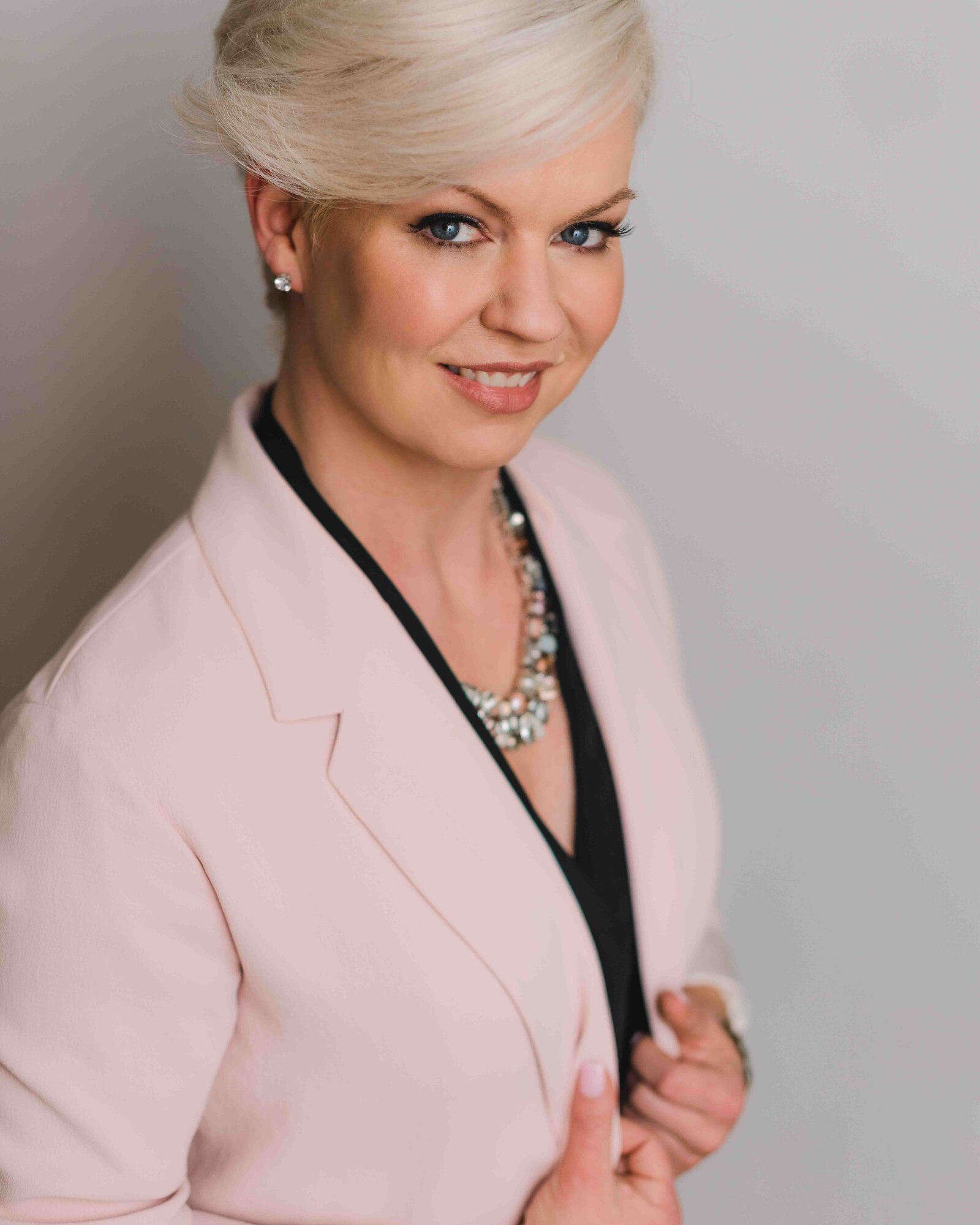 Headshot of a real estate agent with short platinum blonde hair smiles at camera wearing a light pink suit jacket and chunky silver necklace