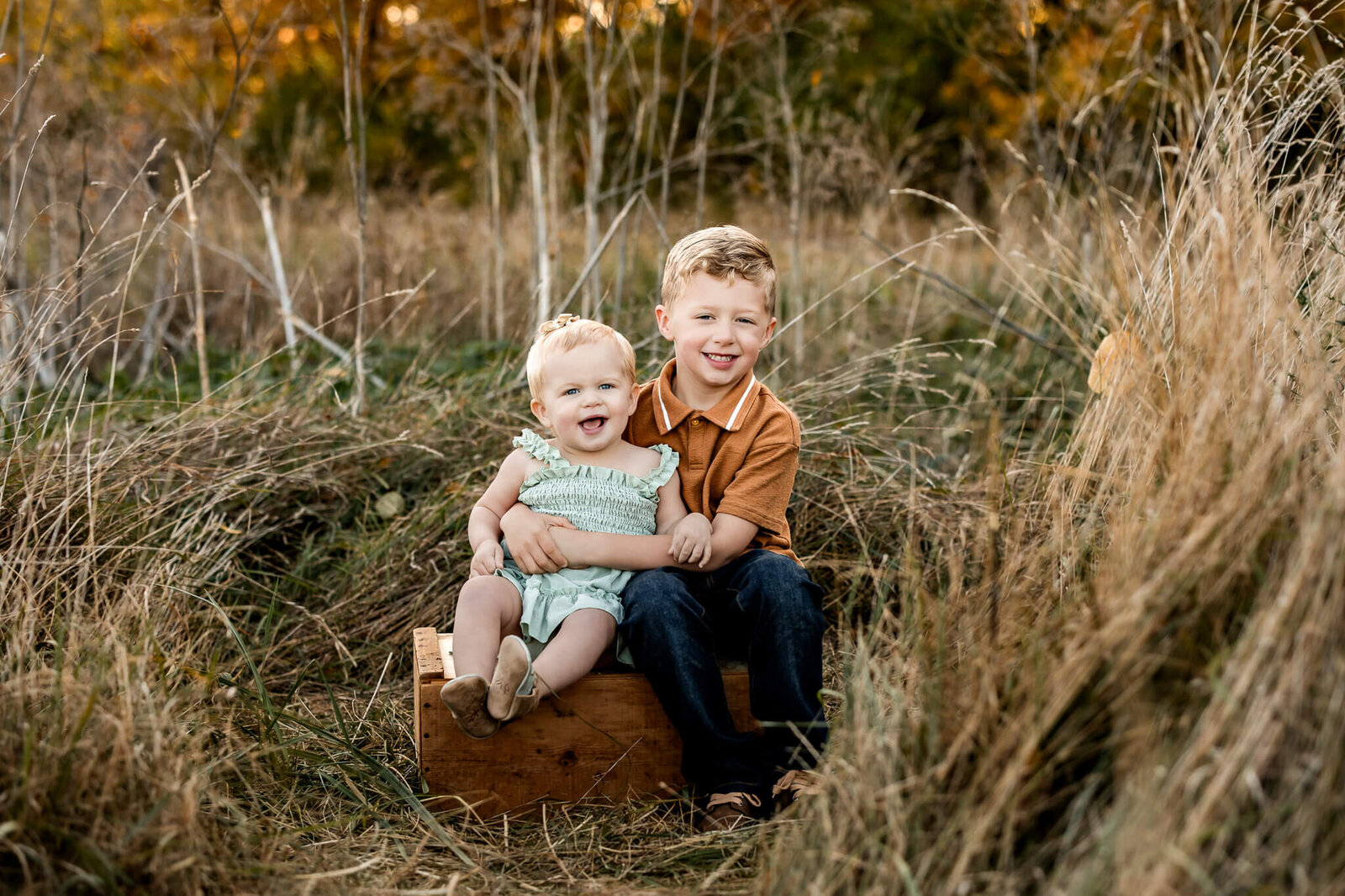 Two young children sitting on a wooden box in a grassy field make for charming outdoor portraits. The toddler on the left is in light green, and the older child wears an orange shirt and jeans. Both are smiling, surrounded by autumn foliage in this picturesque setting captured by a Bloomington family photographer.