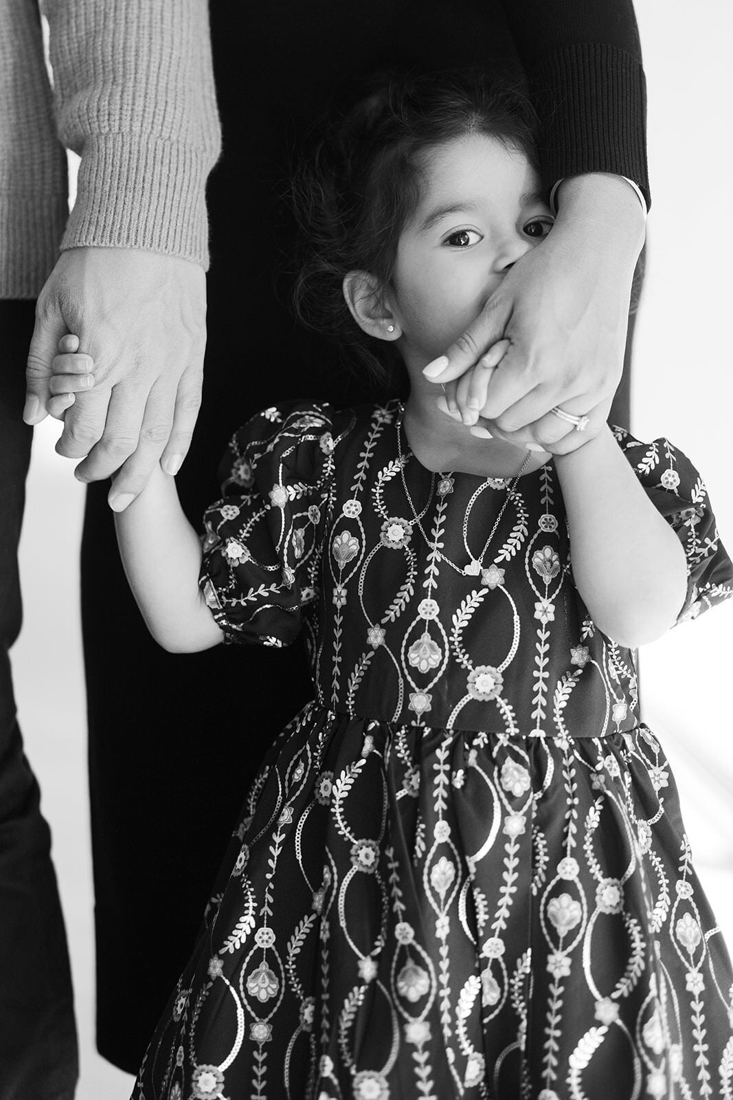 Little girl holding her parents’ hands, her face framed by their arms in an intimate family portrait.