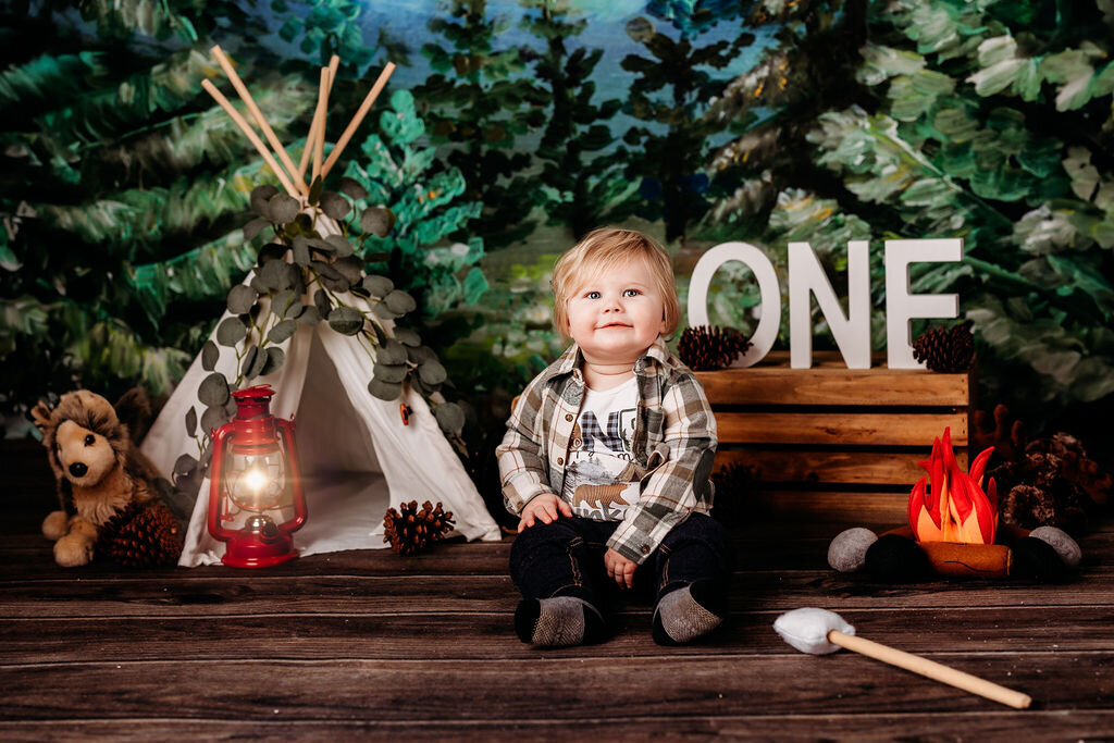 Amongst a custom camping themed backdrop, a baby smiles joyfully for the camera