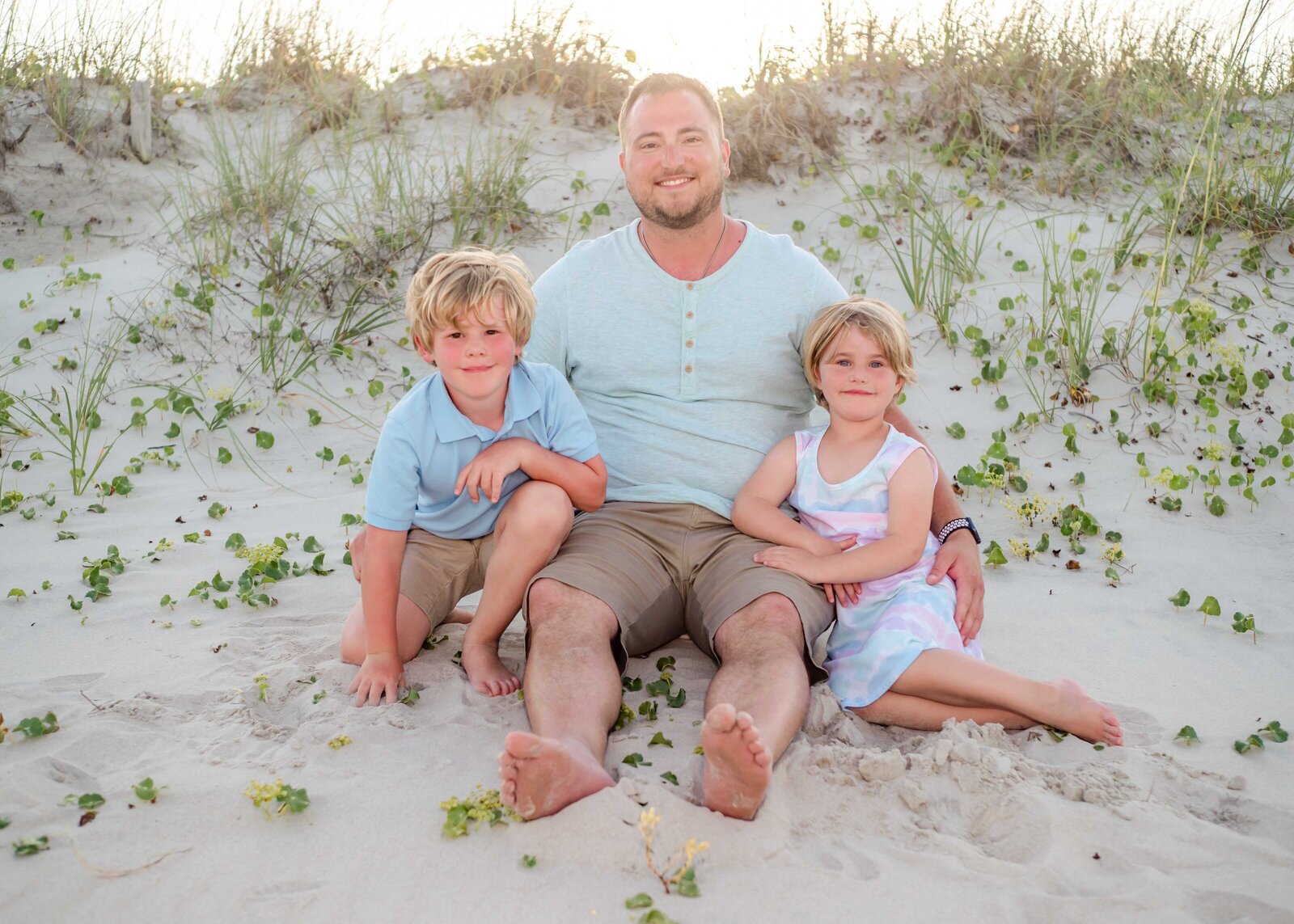 A father sits on the sandy dunes with his two young children at a Wilmington beach, surrounded by greenery. The relaxed and intimate setting is perfect for capturing affordable family photography in Wilmington, NC, focusing on cherished moments.