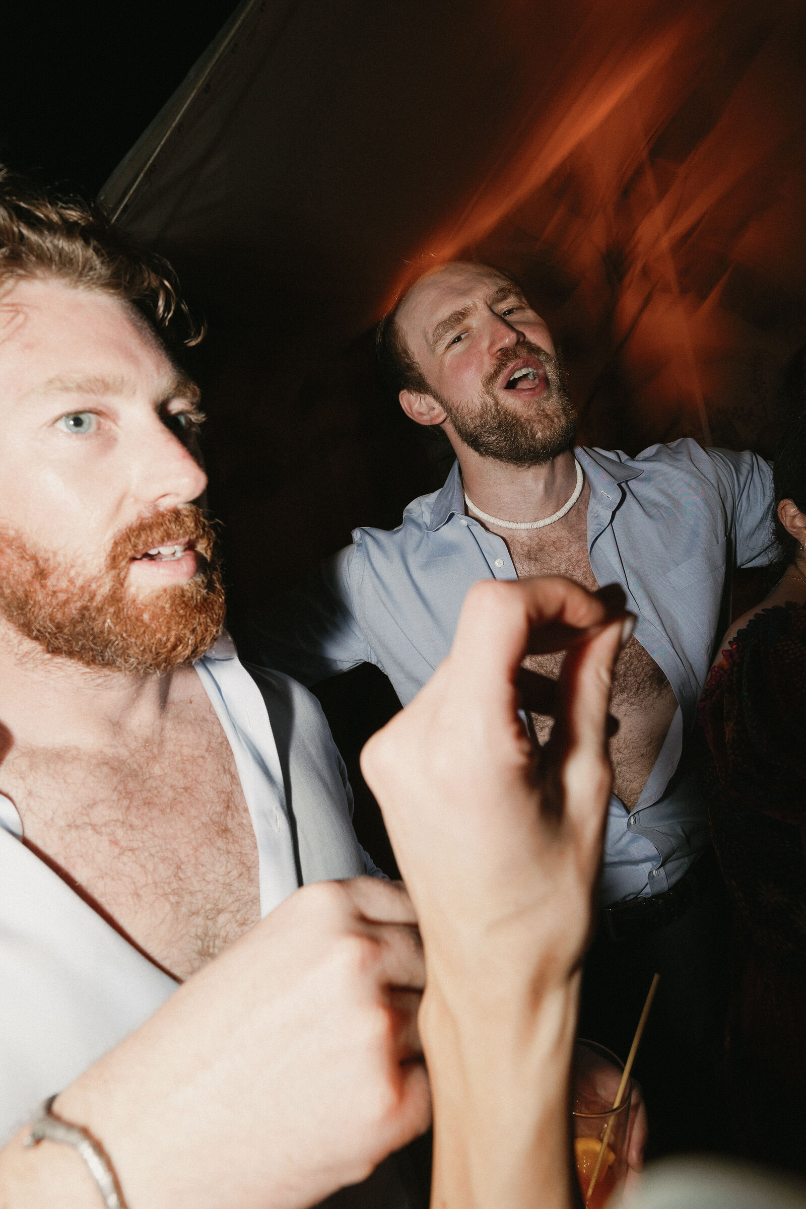 Direct flash photo of two male wedding guests on dance floor with female hand snapping in the foreground