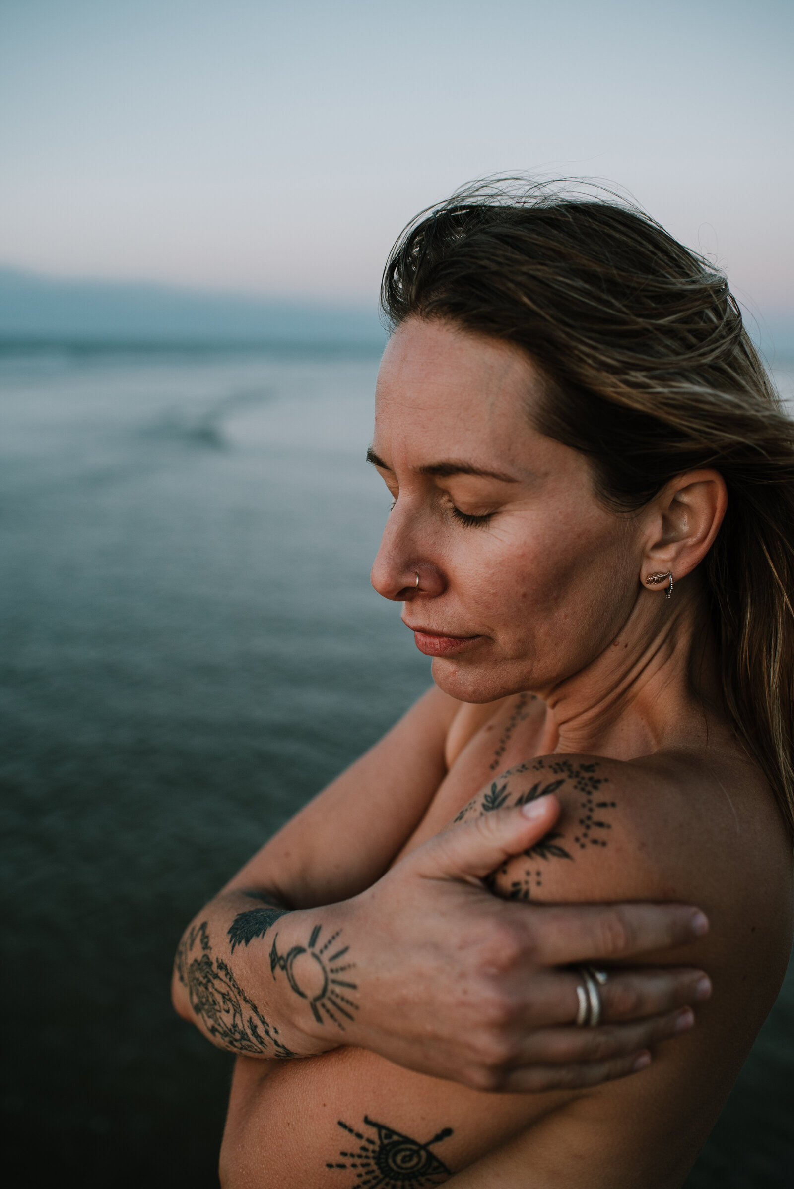 woman is hugging herself close, stood in the blue ocean, her skin is bare and she is covered in tattoos. she looks calm and safe