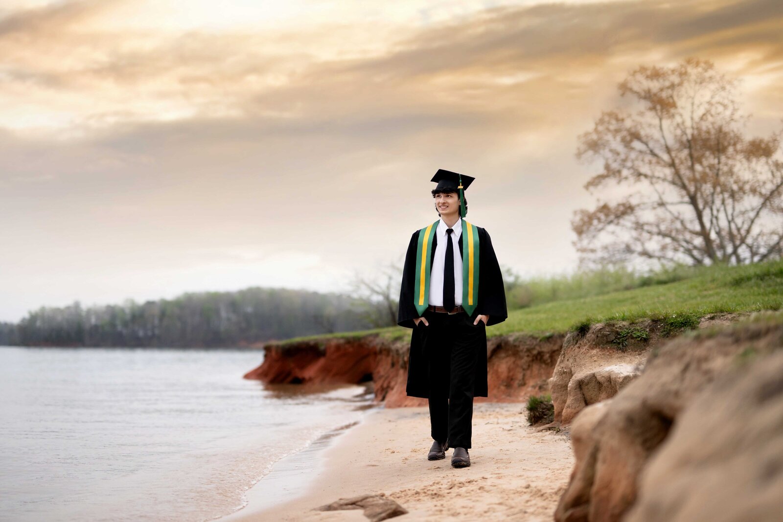 Graduation photo of boy in cap and gown, walking on lake and looking into the sunset. Senior graduation photos taken by Jenni Nikki Photography in Atlanta, GA