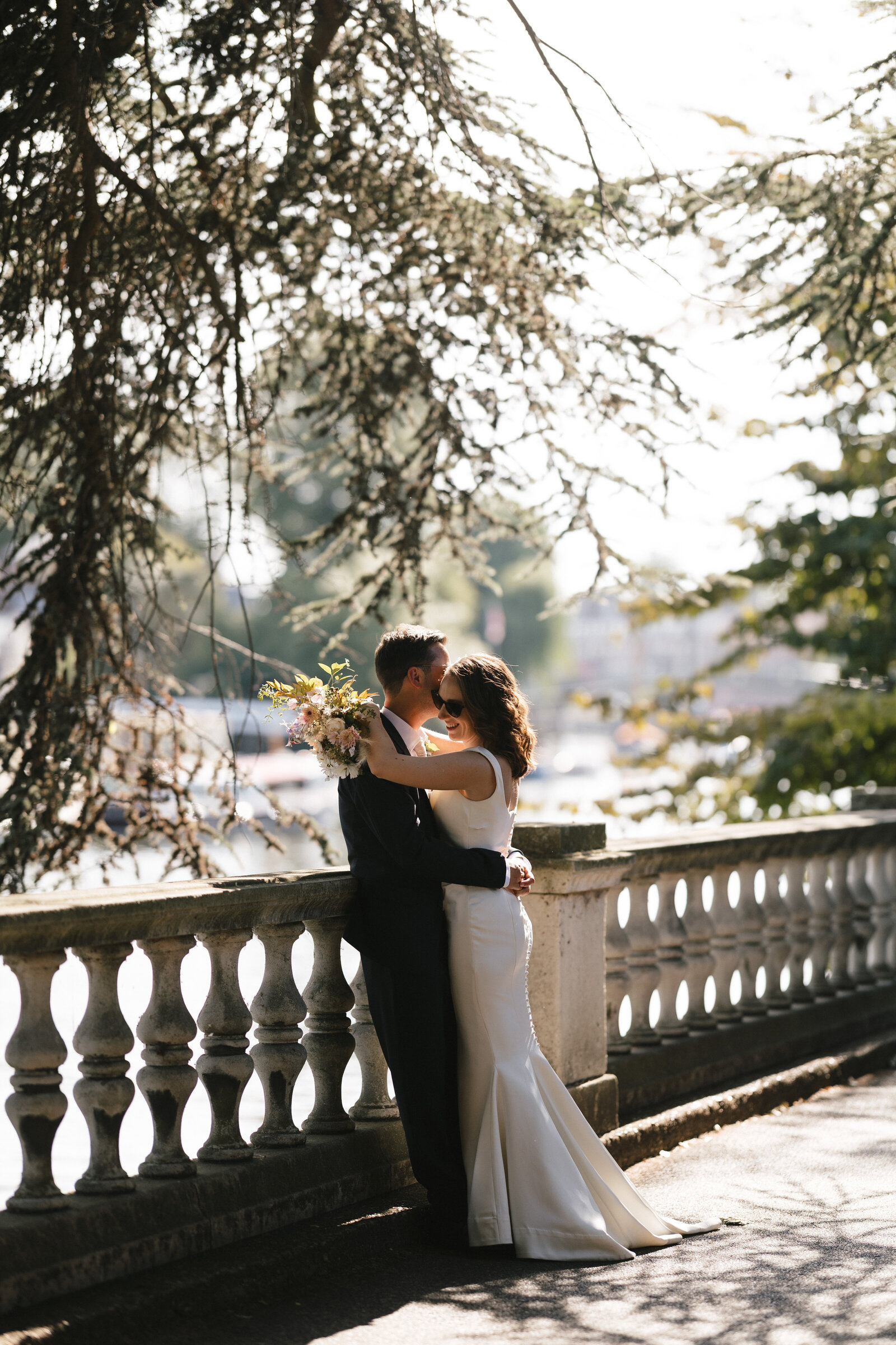 A bride and groom embrace and smile at each other outdoors during their London wedding, with the bride holding a bouquet of flowers. Sunlight filters through the trees, creating a romantic, natural setting by the water
