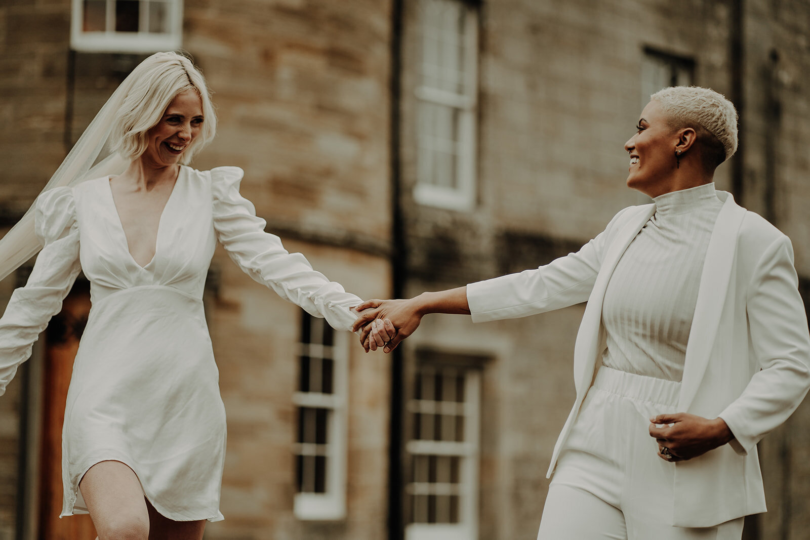 two brides in white outfits holding hands and smiling as they look into each other eyes alternative wedding photographer scotland