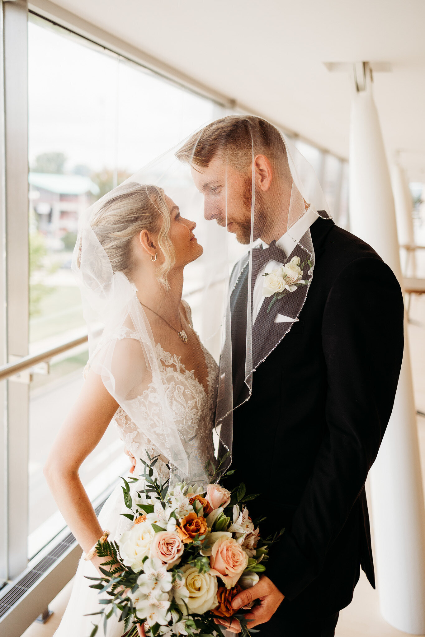 Both under her veil, a bride and groom gaze into each others eyes surrounded by large windows and natural light 