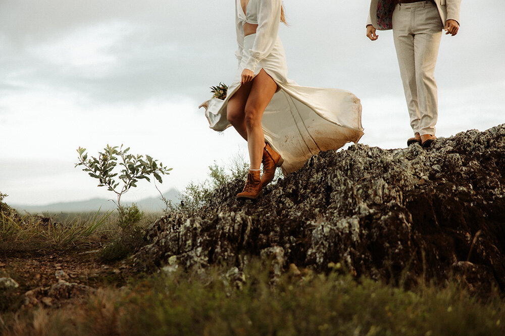 Bride lifting her wedding dress while walking across rugged cliffs during an adventurous elopement shoot, captured by Sunshine Coast elopement photographer.