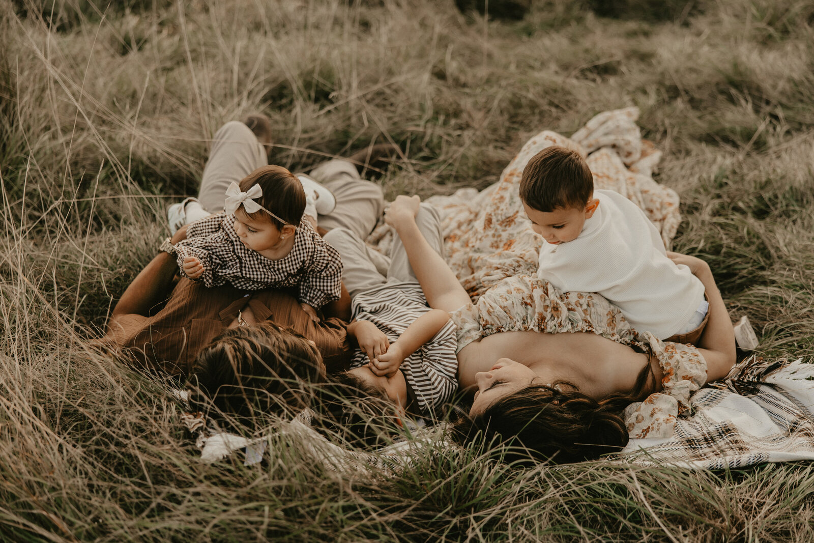 A family of three enjoys a golden hour walk along a scenic dirt path in Morris County, New Jersey. The warm sunset light highlights the tall grass and bare trees, creating a serene and picturesque backdrop. The mother wears a flowing floral dress, the father is dressed in neutral tones, and their young child is bundled in a cozy jacket. 