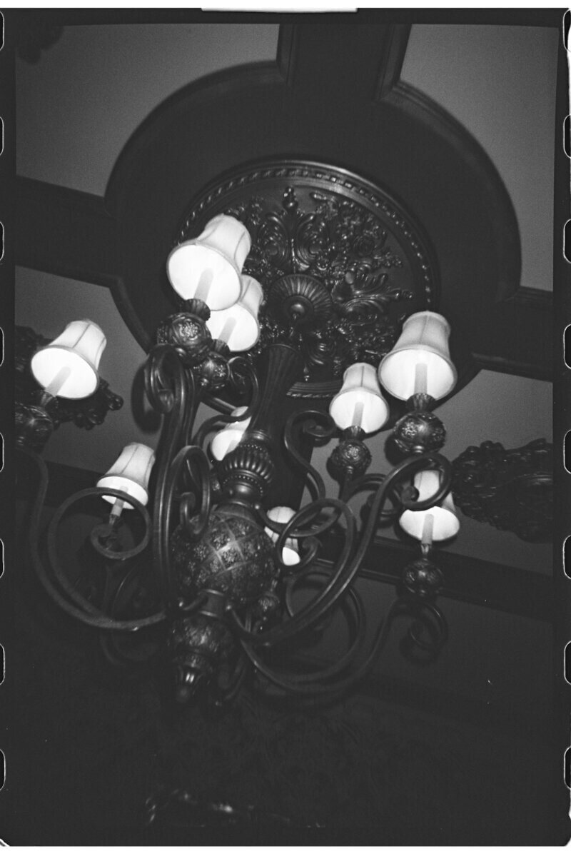 Black and white photo of an ornate chandelier with multiple light bulbs, featuring intricate metalwork and decorative detailing, viewed from below against a decorative ceiling.