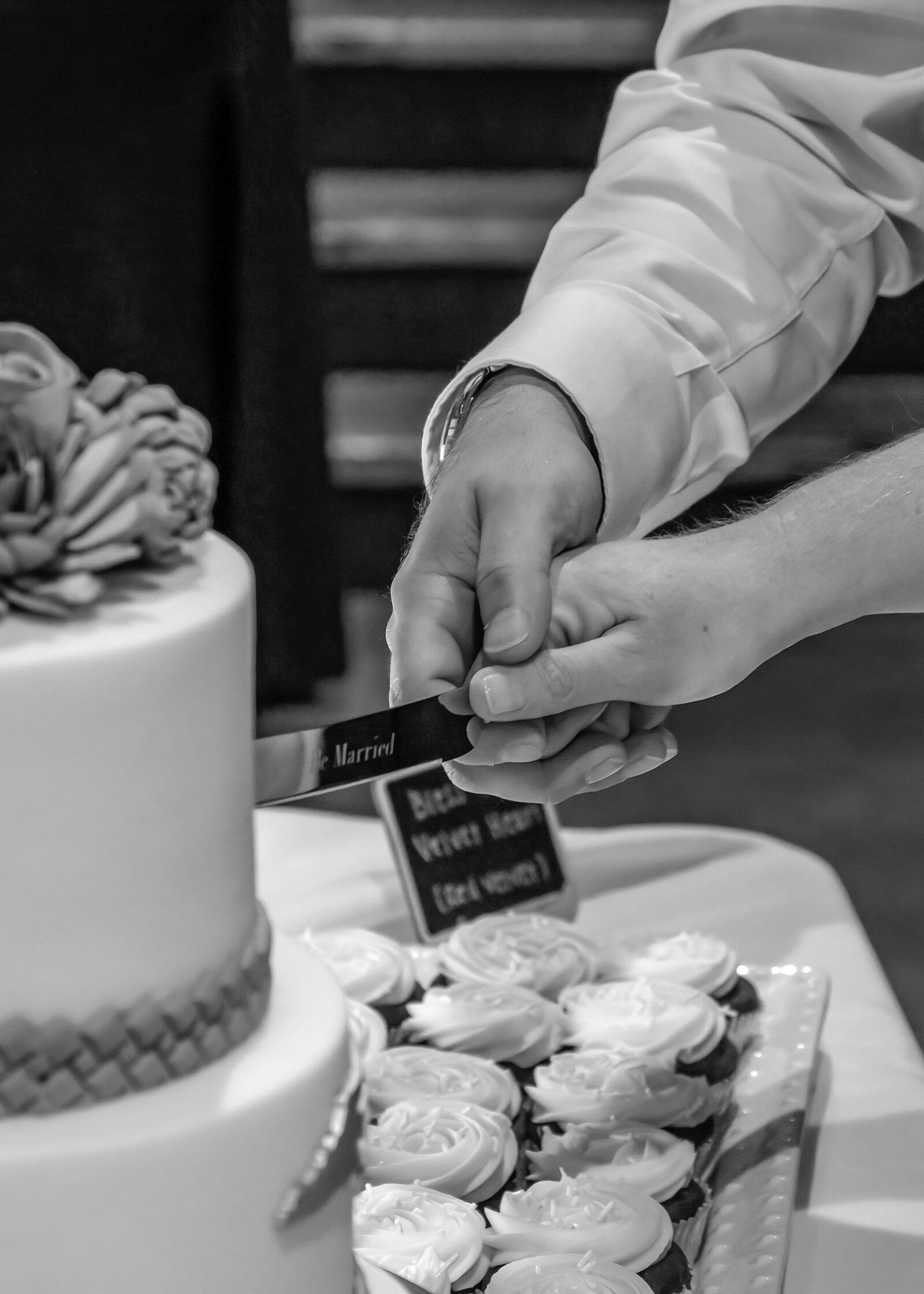Black and white close-up of the bride and groom cutting their elegant wedding cake, surrounded by cupcakes, captured during a Wrightsville Beach, NC wedding, highlighting timeless wedding traditions.
