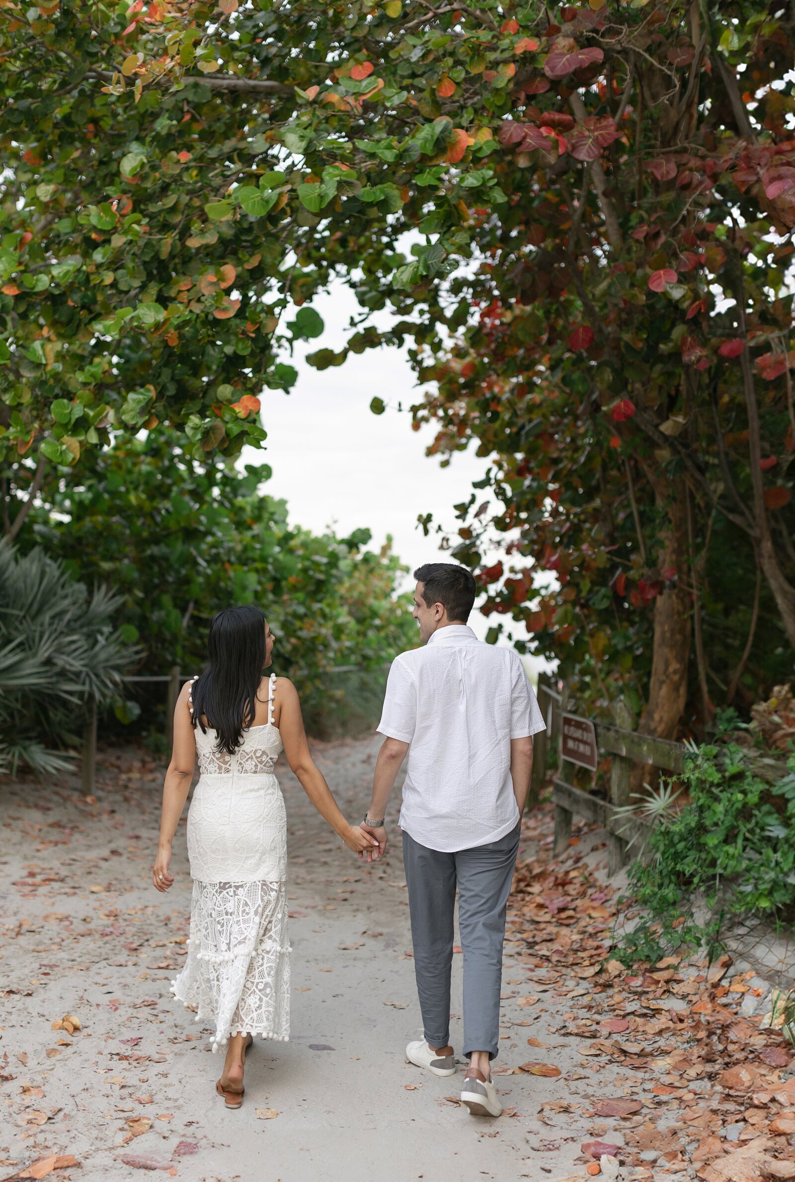 Couple walk hand in hand towards the beach with green trees surrounding them.