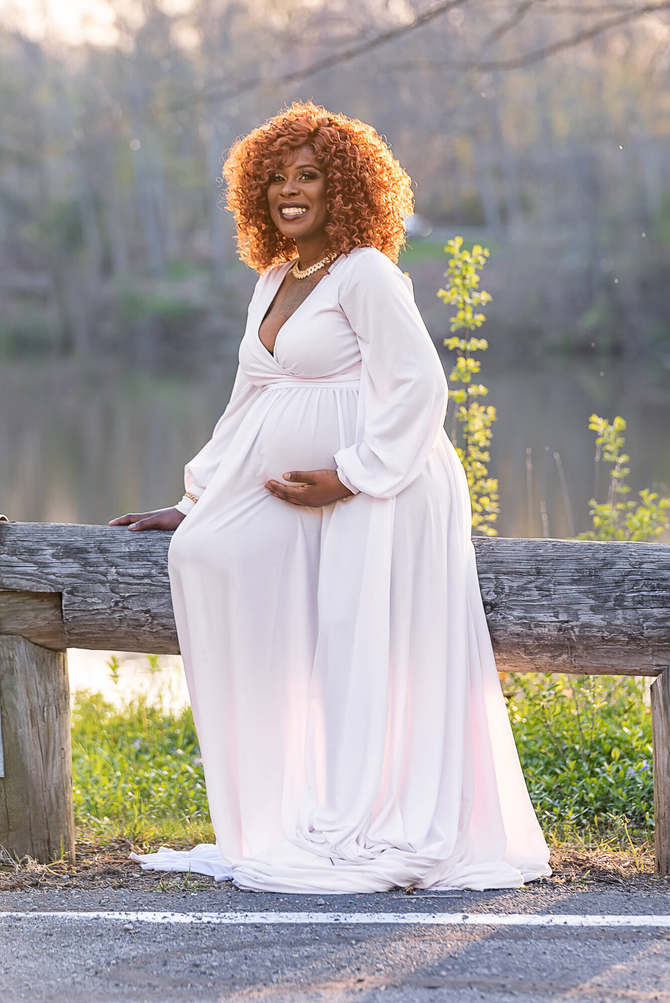 A pregnant woman sits on a wooden fence near a peaceful lake at sunset, wearing a flowing white gown. The golden light adds a dreamy glow to the scene.
