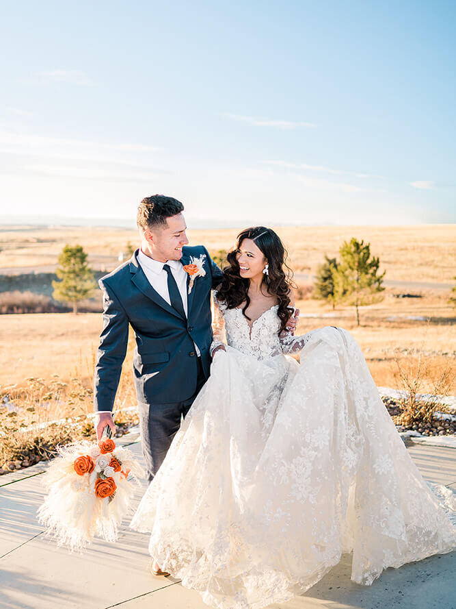 Groom in a navy suit holds his brunette bride's florals while she holds the skirt of her lace gown