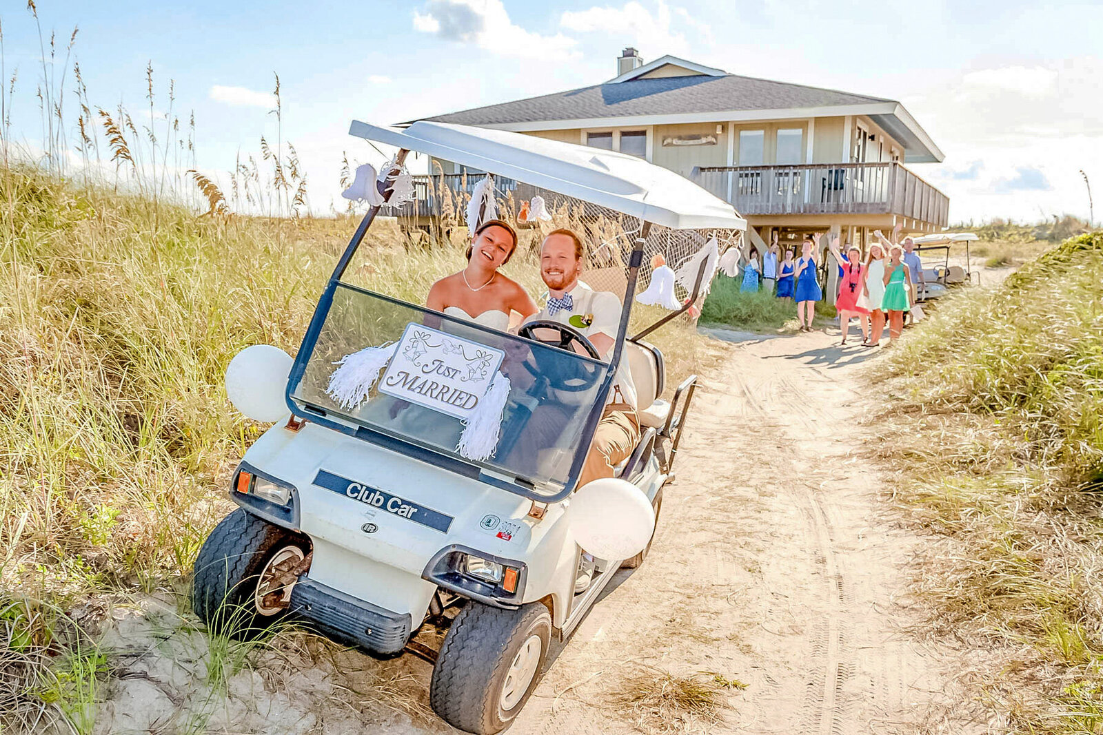 Newlyweds joyfully driving a decorated golf cart down a sandy path after their beach wedding ceremony on Bald Head Island in North Carolina.