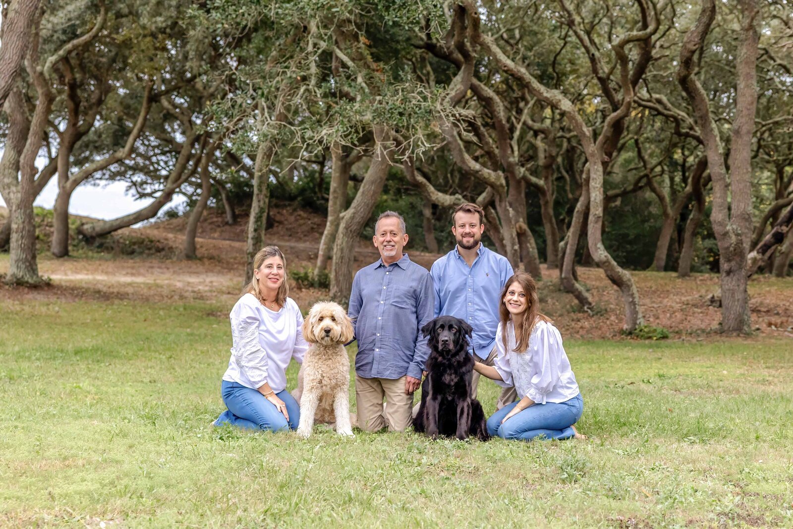A family poses in a wooded park with two dogs, surrounded by lush greenery. Dressed in casual blue and white outfits, they enjoy a peaceful outdoor setting, perfect for professional family photography in NC, highlighting family and pets.