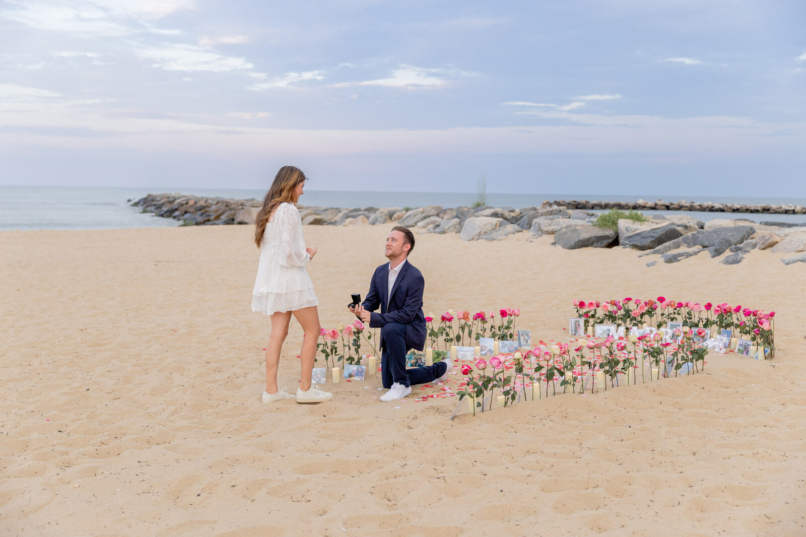 Guy proposing to his girlfriend on the beach on the coast of Virginia. He is kneeling on a bed of rose petals and roses, creating an aisle in the sand.