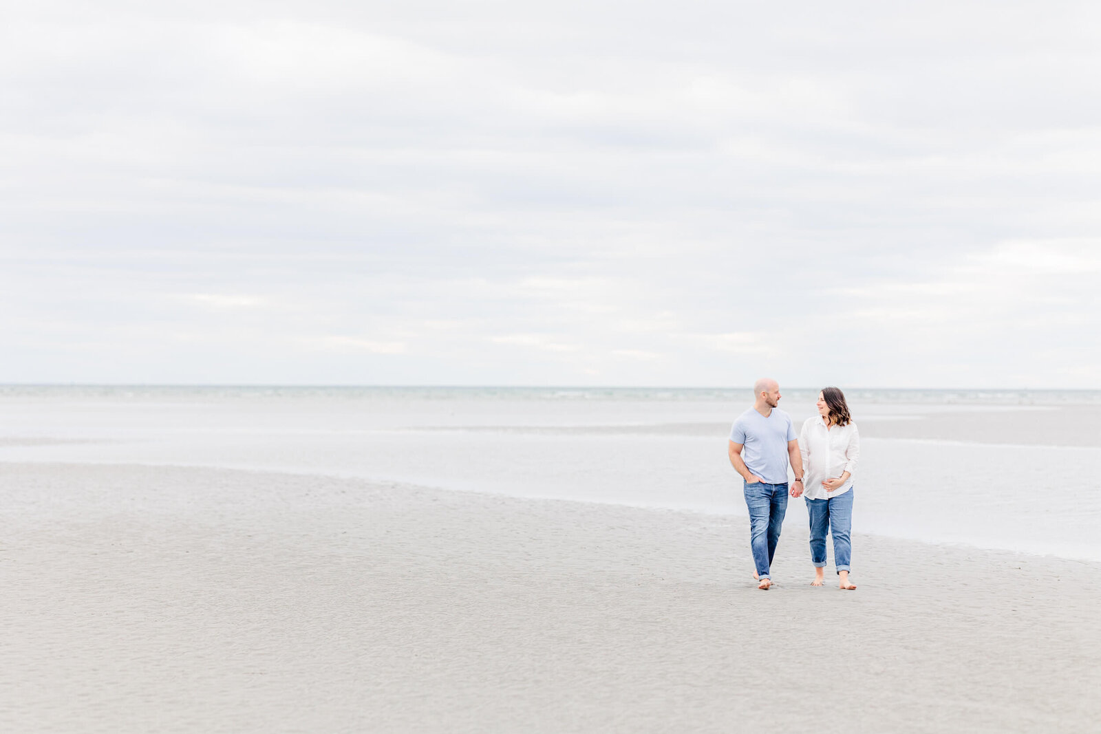 Man and his pregnant wife walk along the ocean on an overcast day