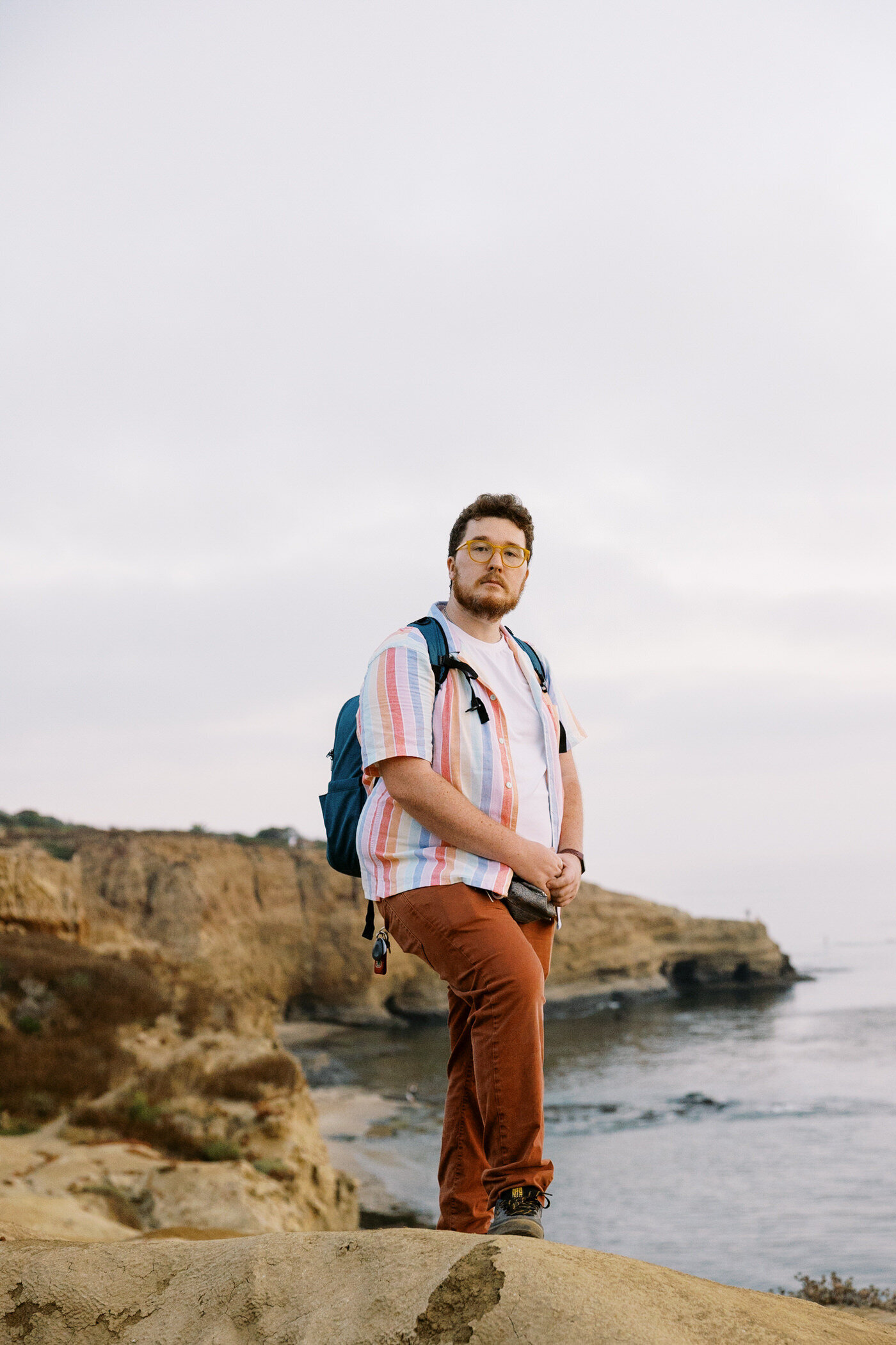 Man wearing terra cotta colored pants with a white shirt and colorful open button down, wearing a camera bag and holding a small camera, poses with one leg up on a rock at sunset cliffs