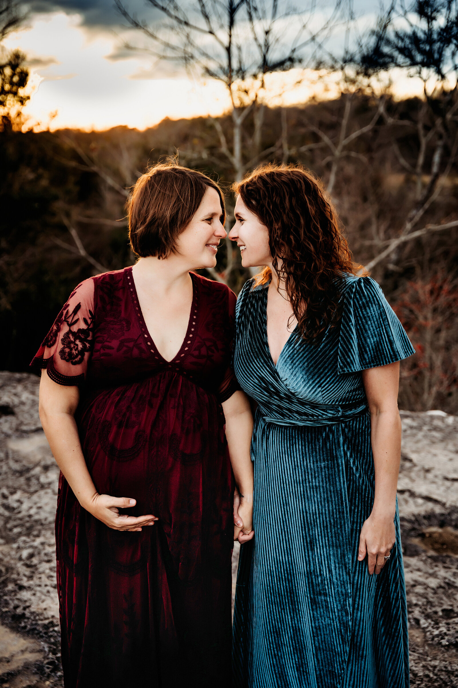 Maternity Photographer, an expectant woman holds hands with her partner, both women smile outside as they gaze at each other