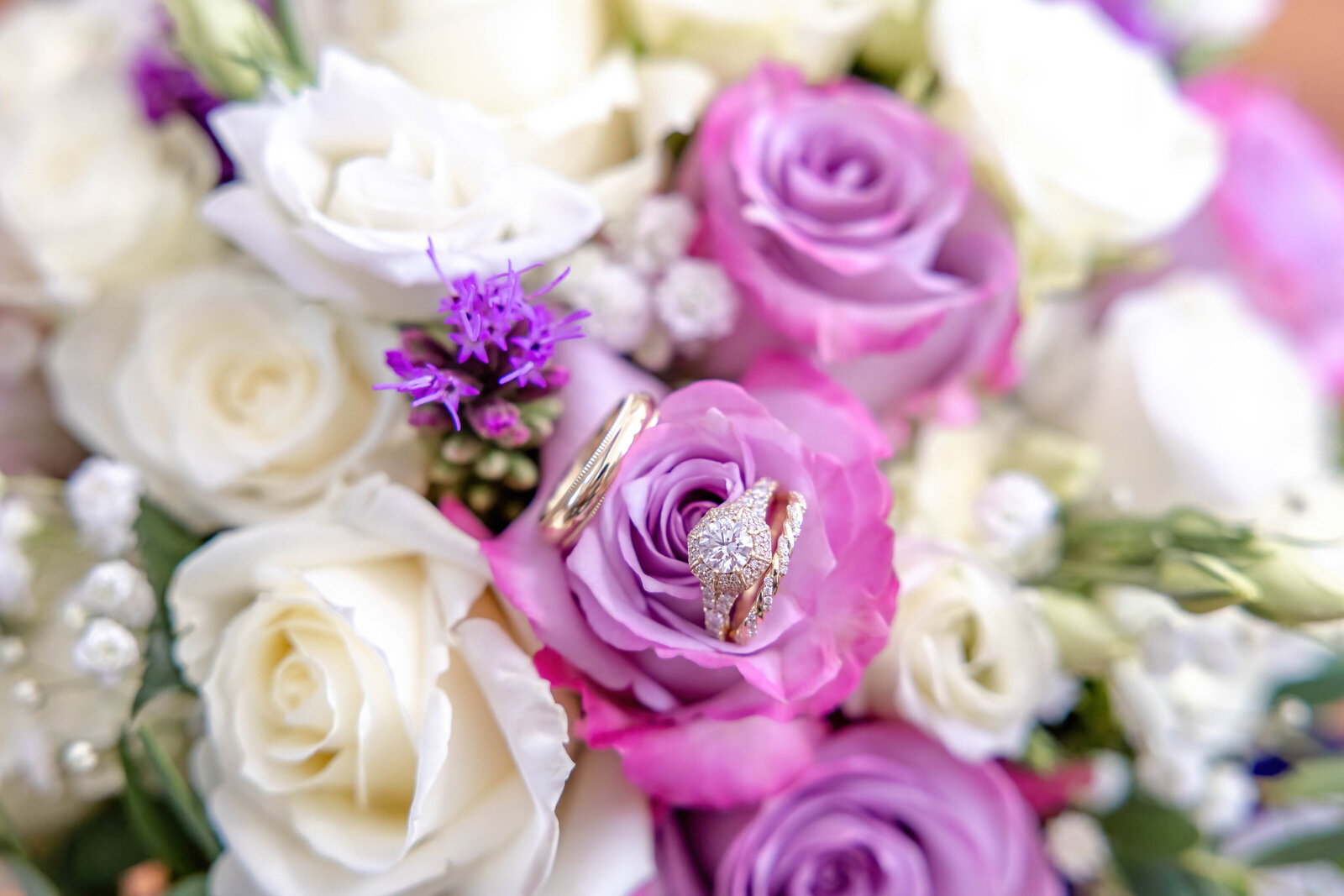 A close-up of wedding rings nestled in a bouquet of purple and white roses with lavender accents, captured during a Wrightsville Beach, NC wedding, showcasing elegant and timeless wedding details.