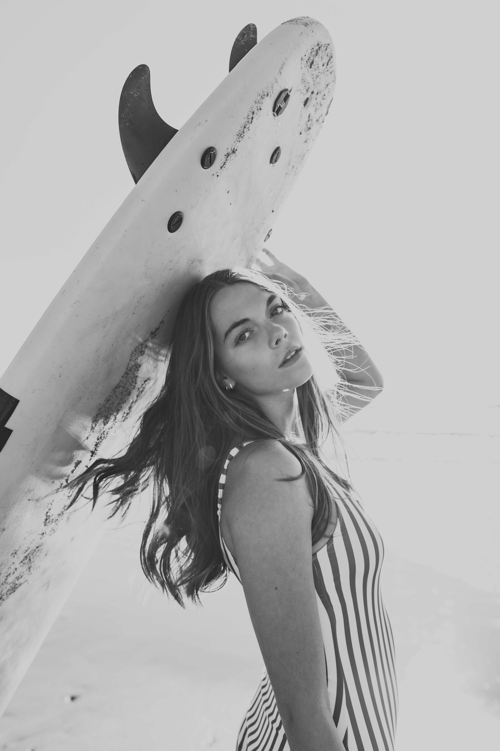 black and white picture of brunette girl holding a surf board wearing a striped one piece swimsuit