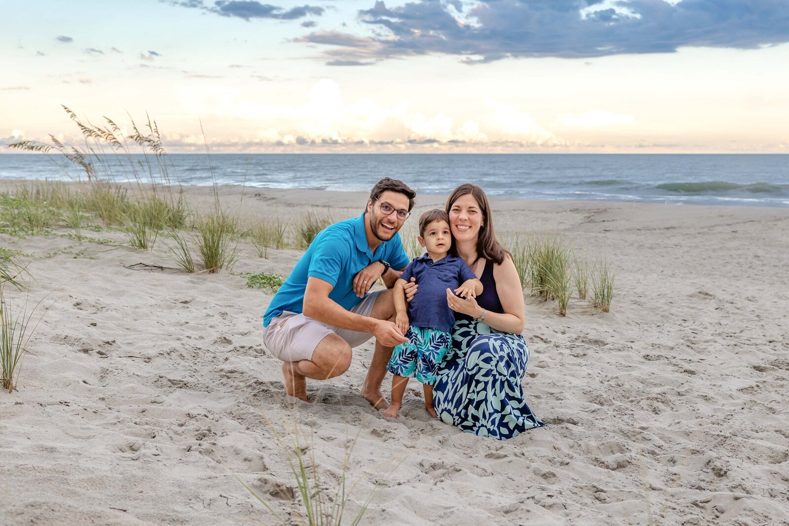 A small family kneels on the beach at dusk, surrounded by sea grass and a calm ocean. Dressed in blue attire, they share a joyful moment, perfect for artistic family photography in Wilmington, NC, highlighting peaceful family interactions.