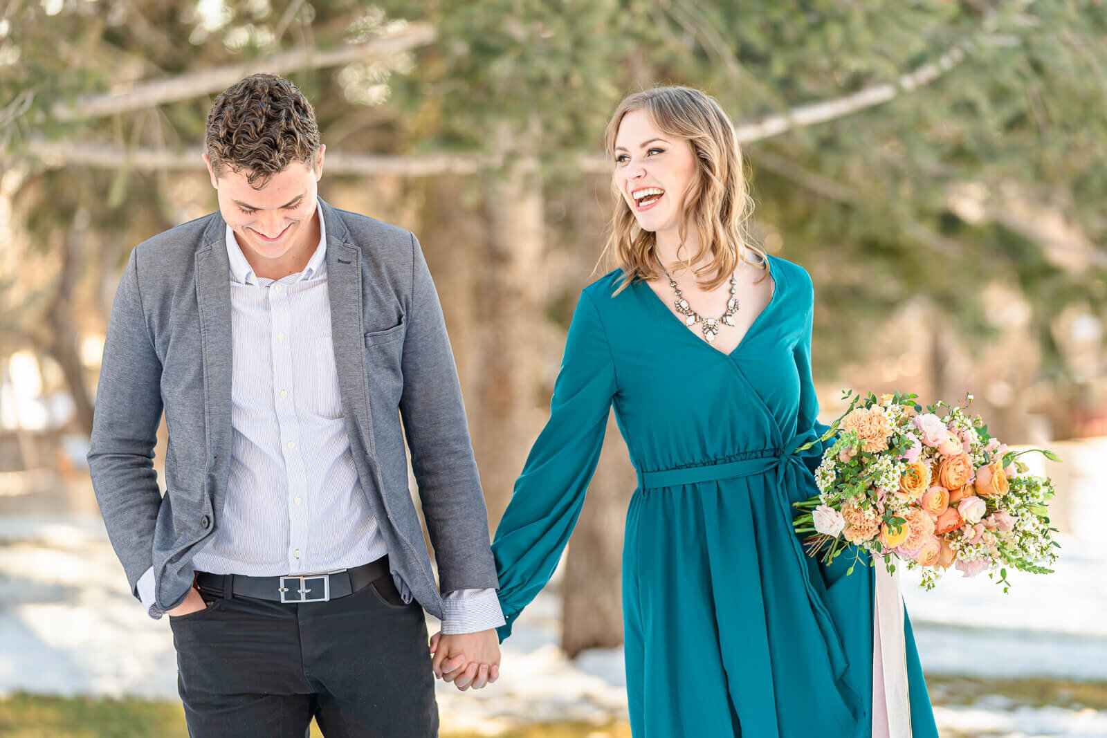 Utah engagement photography of a man in a white button-up shirt and a gray blazer holding hands with his fiancee wearing a long sleeved dark teal dress, holding a bouquet of orange and pink roses while walking in a park in Salt Lake City