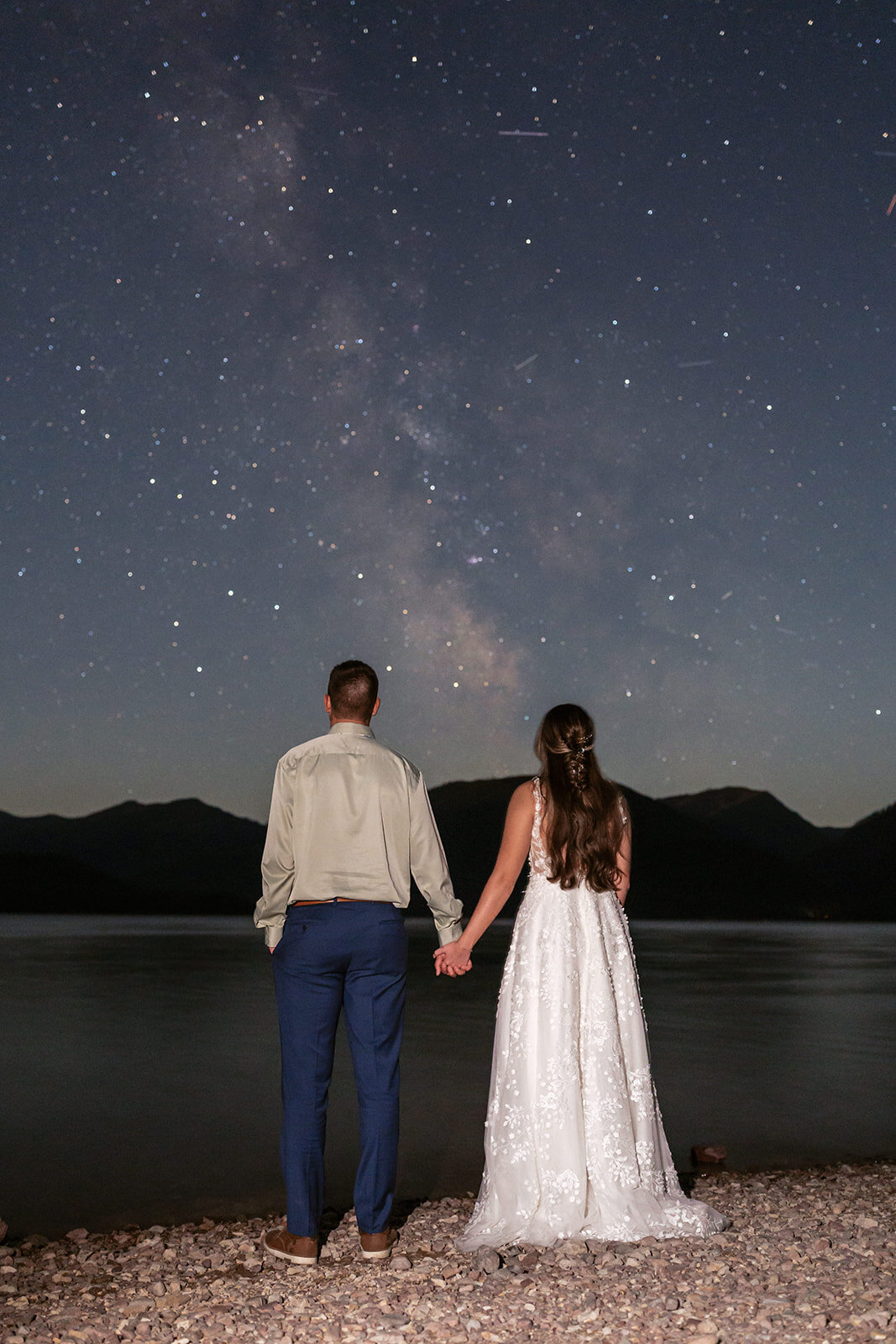 A couple holds hands beneath a star-filled sky reflected on Lake McDonald in Glacier National Park, captured by Sydney Breann Photography during their romantic night elopement.