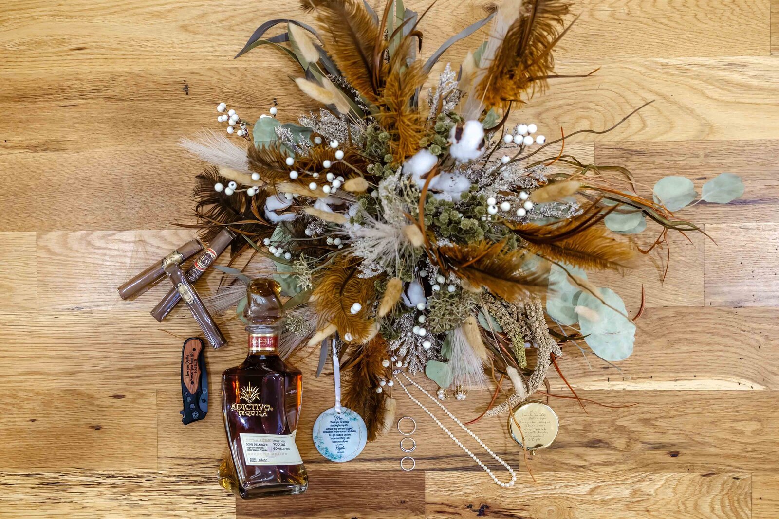 A rustic bridal flat lay featuring a dried floral bouquet, a bottle of bourbon, and personalized wedding charms, captured on wooden flooring during a Asheville, NC wedding, showcasing creative and unique wedding details.