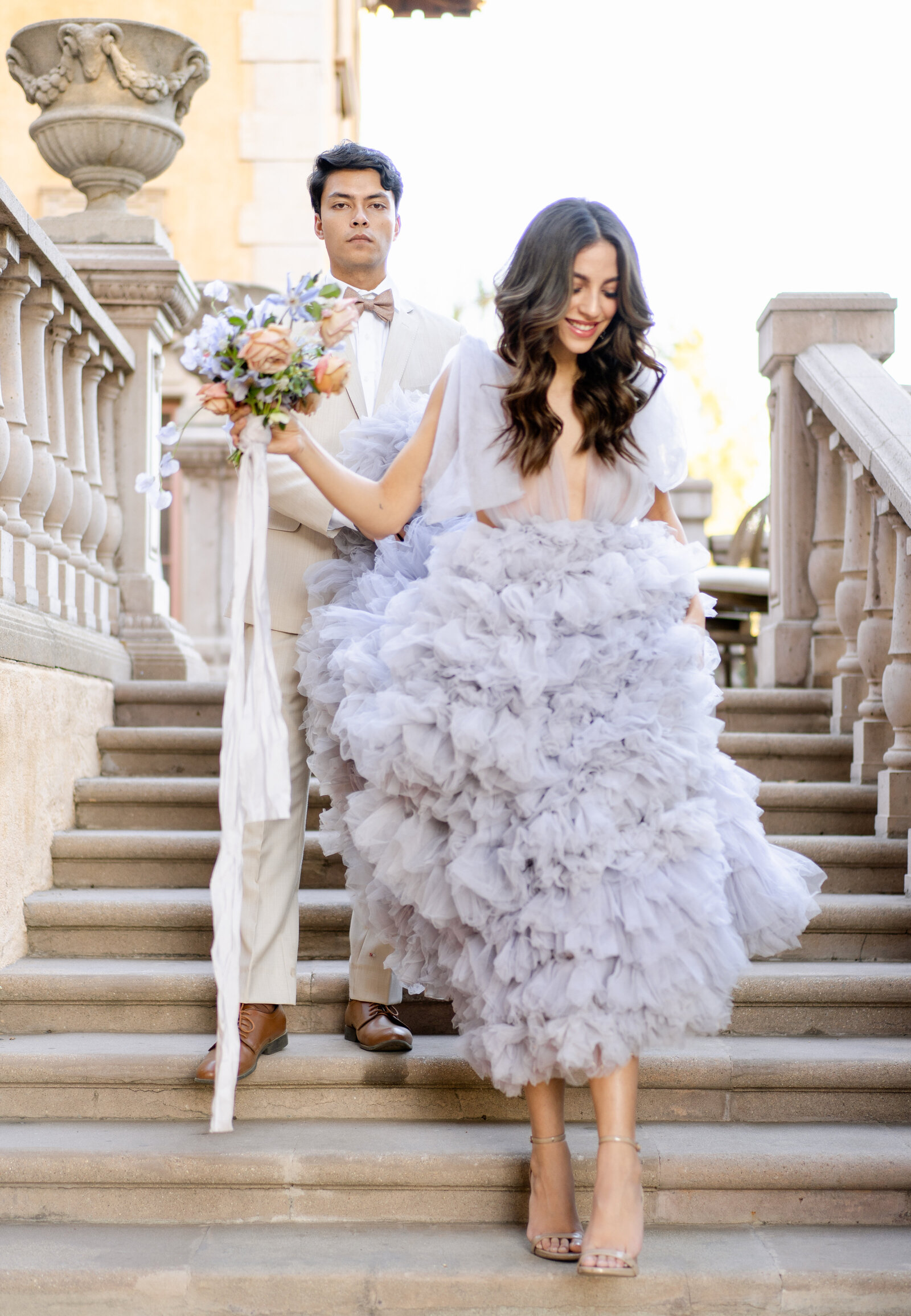 A bride and groom in a lavender wedding gown and cream suit standing on a concrete staircase outdoors.