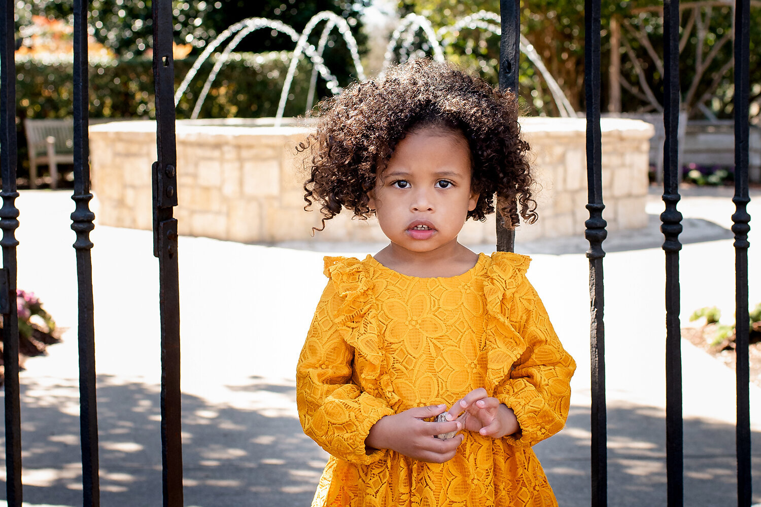 An adorable 3 year old girl wearing a yellow dress standing in front of an iron gate.