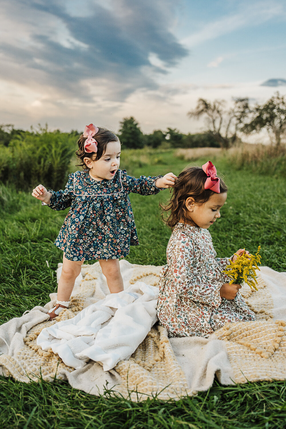 toddler girl pulls sisters hair as she sits on blanket with a bouquet of wildflowers