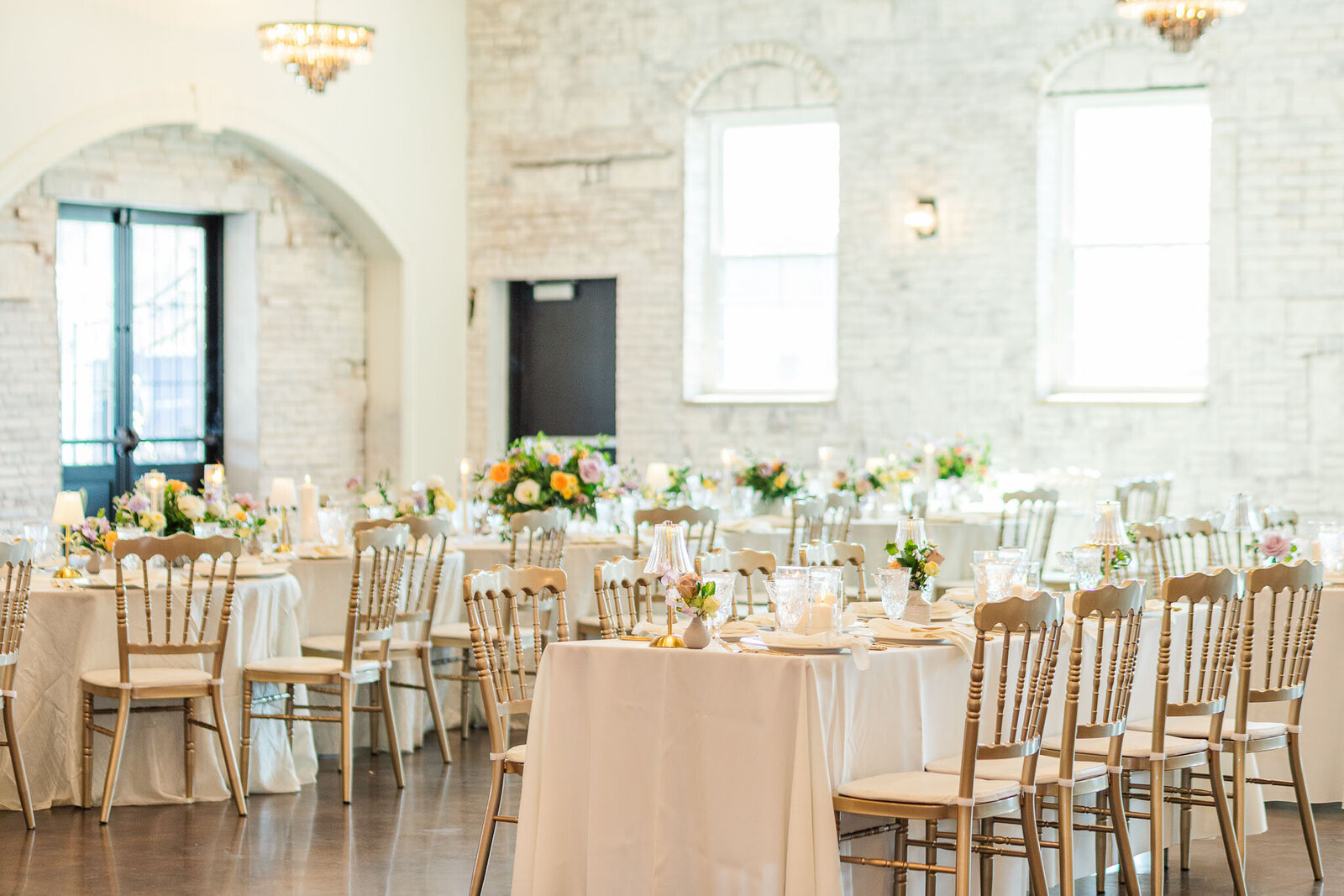 A bright and elegant wedding reception room at Grbic Events in St. Louis, featuring white brick walls, round tables with white linens and pastel floral centerpieces, and Chiavari chairs arranged for guests.
