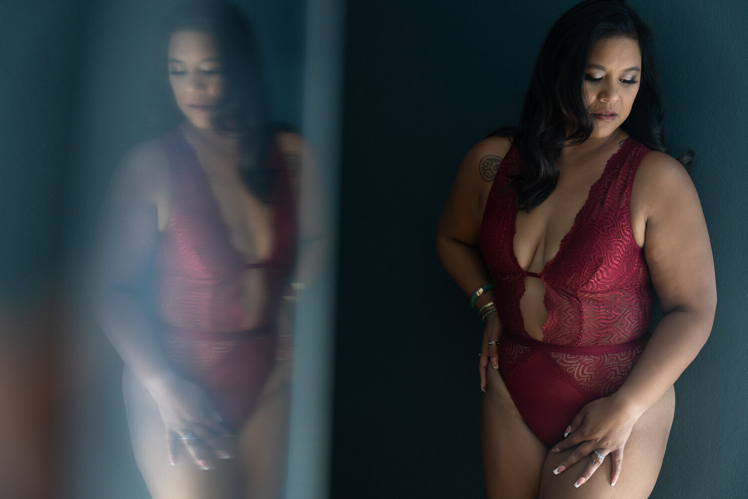 A woman stands against a dark blue backdrop, draped in a dark red lace lingerie bodysuit. Her pose is composed, with one hand resting on her hip, and the other on her thigh, her gaze thoughtfully directed towards the floor. The image captures her figure on the right side, with a mirrored reflection on the left, creating a dramatic and alluring visual symmetry.