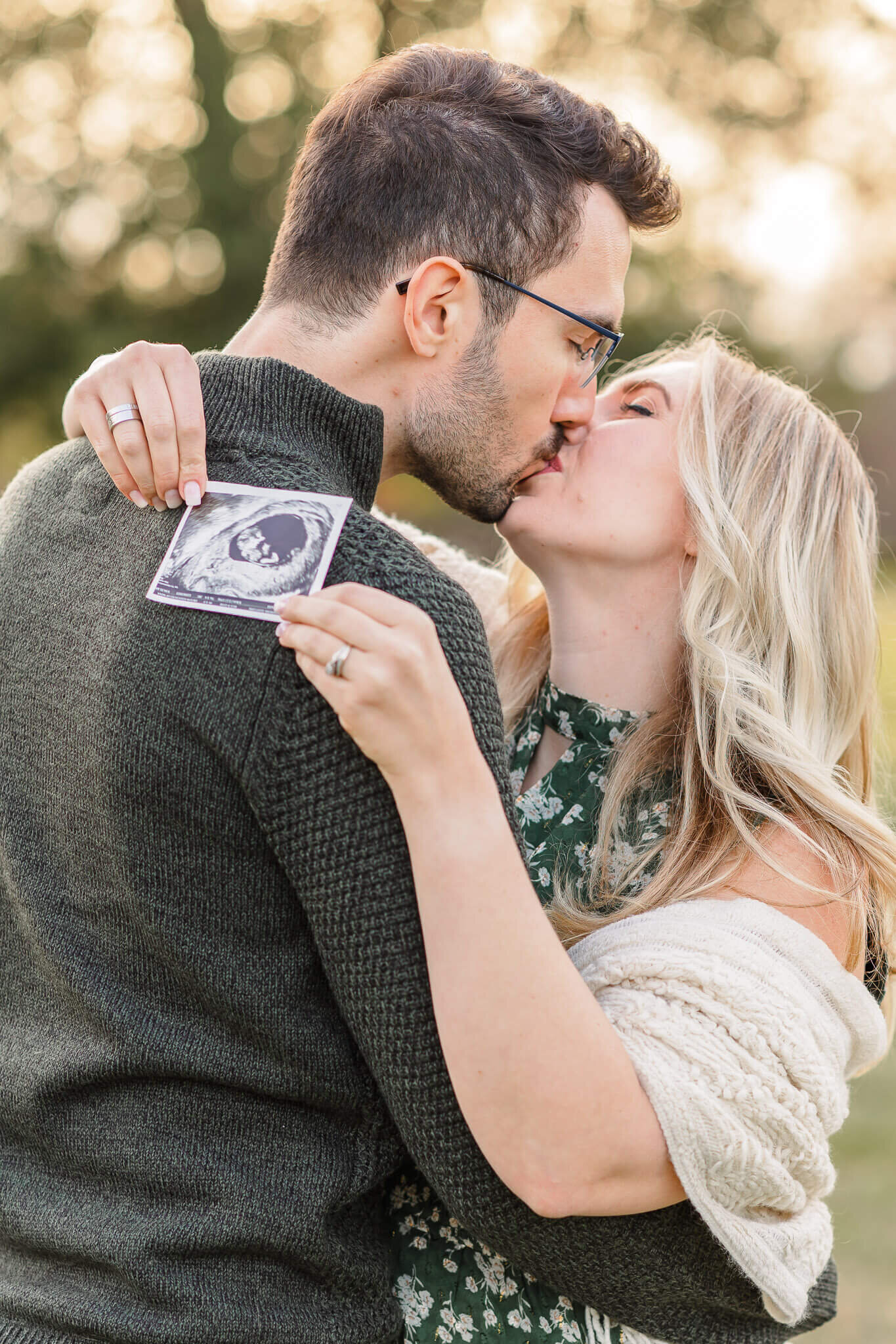 A mom and dad kissing while the mom holds an ultrasound over the dad's shoulder.